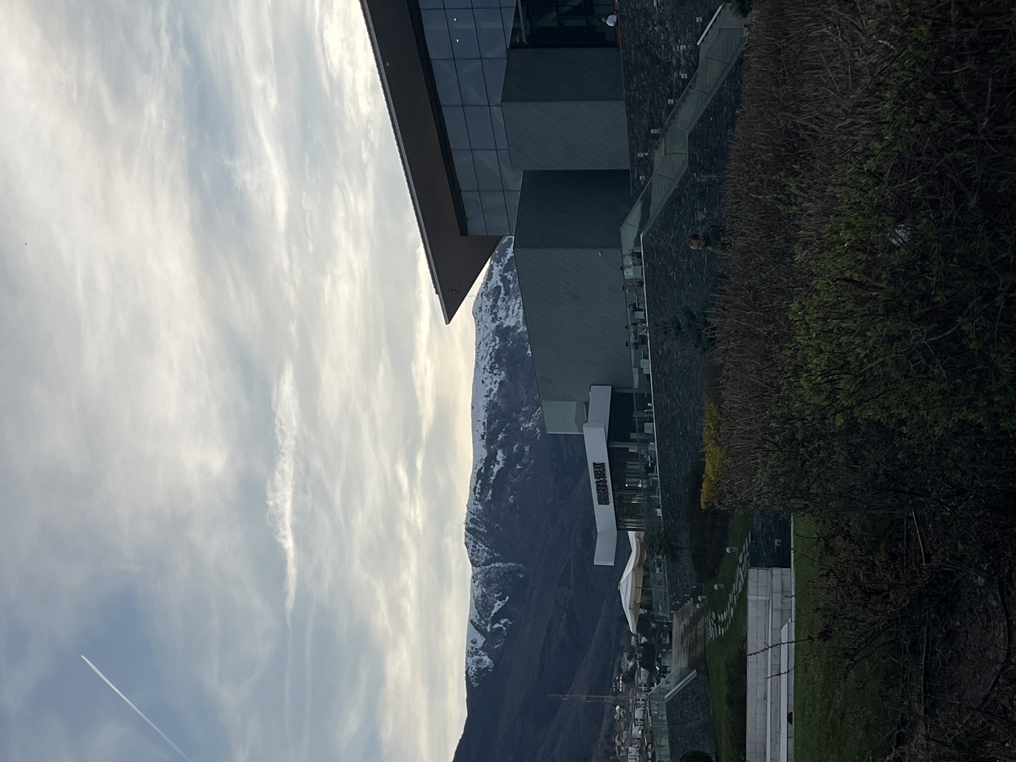 The image shows a cityscape with a modern building, a mountain range, and cloudy sky. The building has a geometric design with glass and dark panels. In front of the building is a public area with trees, grass, and stone walkways. People can be seen walking or sitting on the building's terrace.
In the background, a mountain range rises, its peaks partially covered in snow. The sky is filled with clouds, and there is a contrail of an airplane streaking across the sky.
It is not possible to determine the specific location in Sofia, Bulgaria, without more information.