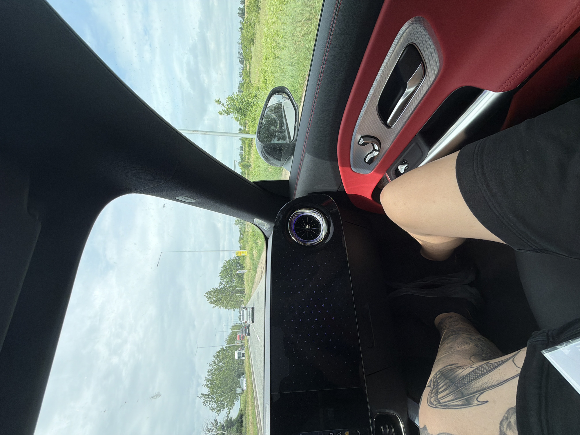 This is a first-person perspective, interior shot from a vehicle driving on a road in Dolno Ablanovo, Bulgaria. The passenger's legs are visible in the foreground, with tattoos on the left leg. The car's interior features red leather accents on the door panel and a digital dashboard display. Outside the car, trees and fields line the road, and other vehicles are visible in the distance. The sky is cloudy.
