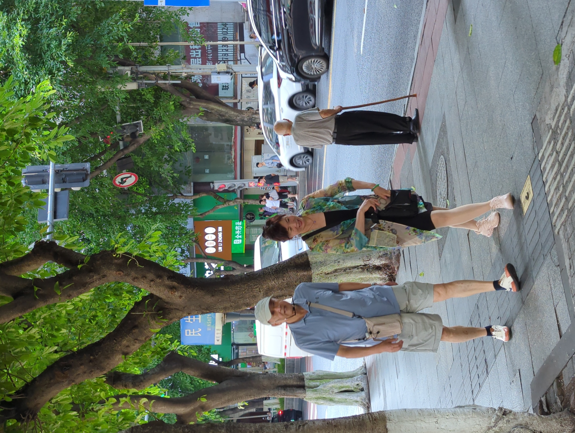 A medium shot captures a street scene in Yuzhong District, China, featuring several individuals and vehicles on a paved sidewalk and adjacent road. In the foreground, a man wearing a light blue polo shirt, grey shorts, and a light-colored baseball cap is walking towards the viewer, with a beige crossbody bag slung over his shoulder. To his right, a woman in a patterned green and black jacket over a black dress is walking in the same direction, looking downwards and holding a dark handbag. Further down the sidewalk, an elderly man in a grey t-shirt and black pants stands with a cane, facing away from the camera and towards the road.
The sidewalk is made of grey paving stones, with some textured areas for tactile paving. Trees with large trunks and green foliage line the left side of the sidewalk, casting shadows. Across the street, a white van is parked, and several cars are in motion, including a black sedan on the right side of the frame. Traffic signals and signs are visible on poles along the street. Buildings with various signs, some in Chinese characters, are present in the background. The overall lighting suggests it is daytime, likely overcast or late afternoon, given the softer shadows.