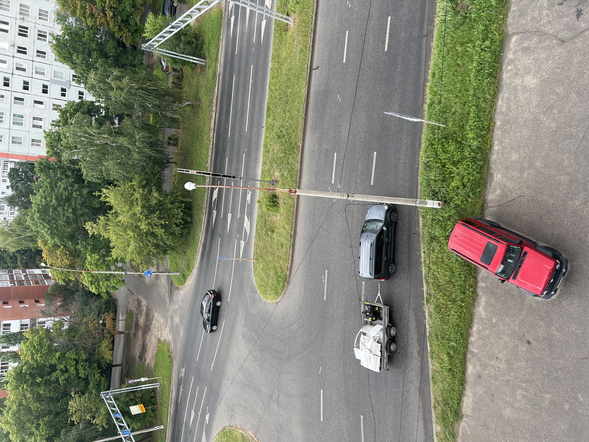 This high-angle, full-shot image, likely taken from an apartment window in Tolyatti, Russia, depicts a segment of a city street during daylight hours. The scene is dominated by a multi-lane asphalt road with white lane markings. On the road, a dark-colored sedan is driving in the left lane, heading away from the viewer. Further ahead, a dark gray SUV is towing a white, enclosed trailer laden with what appears to be construction materials or supplies. The trailer has a dark-colored, possibly yellow, piece of equipment on its tongue.
To the right of the road, a patch of lush green grass lines the asphalt, separating it from a sidewalk and beyond that, residential buildings. The buildings are typical of Soviet-era architecture, with multi-story apartment blocks featuring numerous windows and balconies. Some trees, both deciduous and coniferous, are interspersed throughout the urban landscape, with some showing signs of autumn coloration.
In the foreground, a bright red SUV is parked on a paved area, possibly a street or parking lot, positioned diagonally to the bottom edge of the frame. The red SUV has a sunroof and appears to be in good condition.
Several streetlights and traffic signs are visible along the roadside. A large metal gantry with traffic signs is situated on the left side of the road. A prominent concrete lamppost with electrical wires is positioned near the center of the image, and another traffic sign on a wooden post is visible in the grassy median. The overall impression is a typical urban street scene in a Russian city.