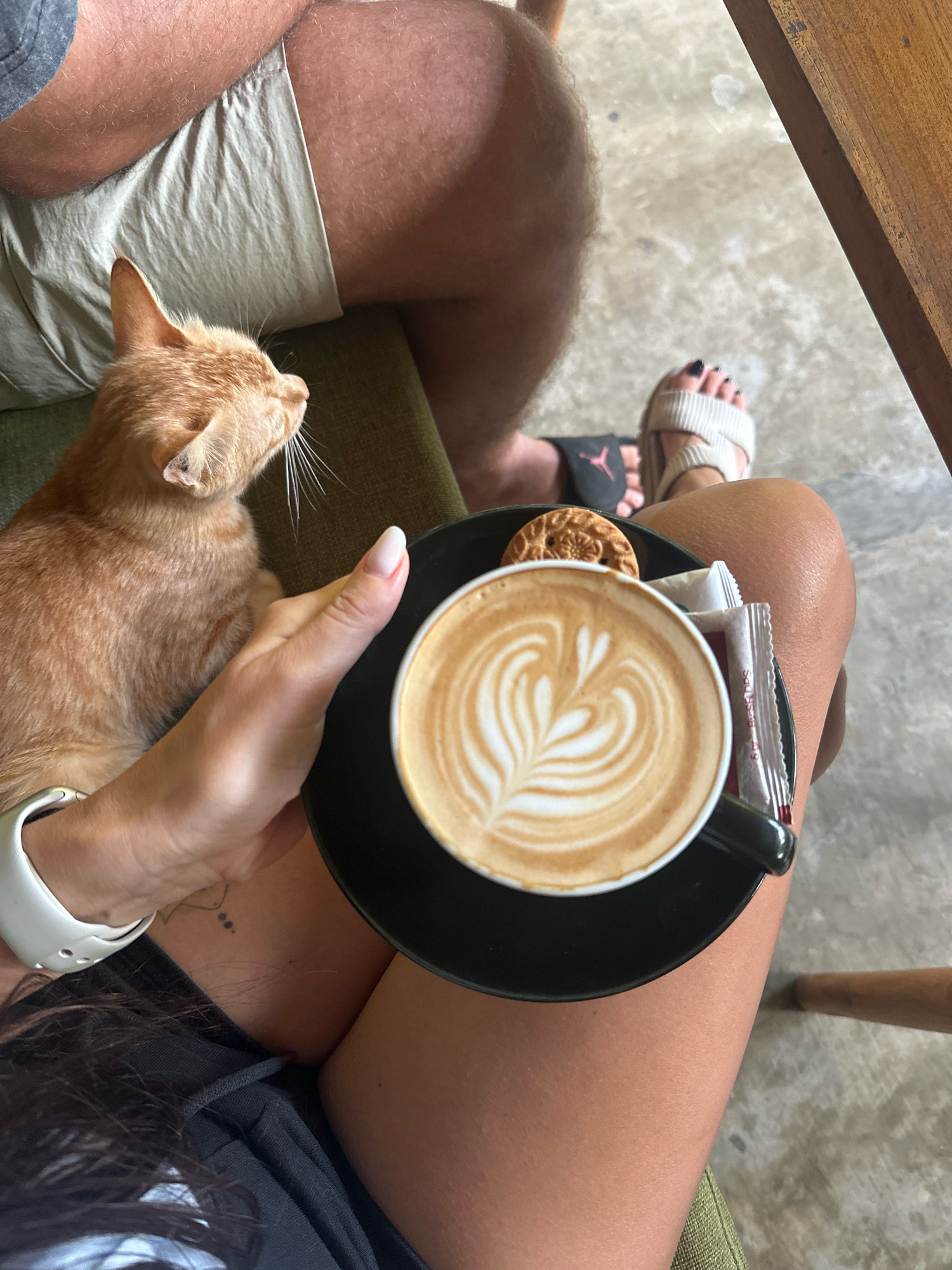 An overhead medium shot captures a woman's hand holding a black saucer with a latte, a cookie, and two sugar packets. The latte has intricate latte art. An orange cat rests on the woman's lap to the left. The woman's legs are visible, wearing a black skirt. To the right, a person wearing sandals sits with their legs crossed. The background shows a concrete floor and a wooden table leg. The setting appears to be an outdoor cafe.