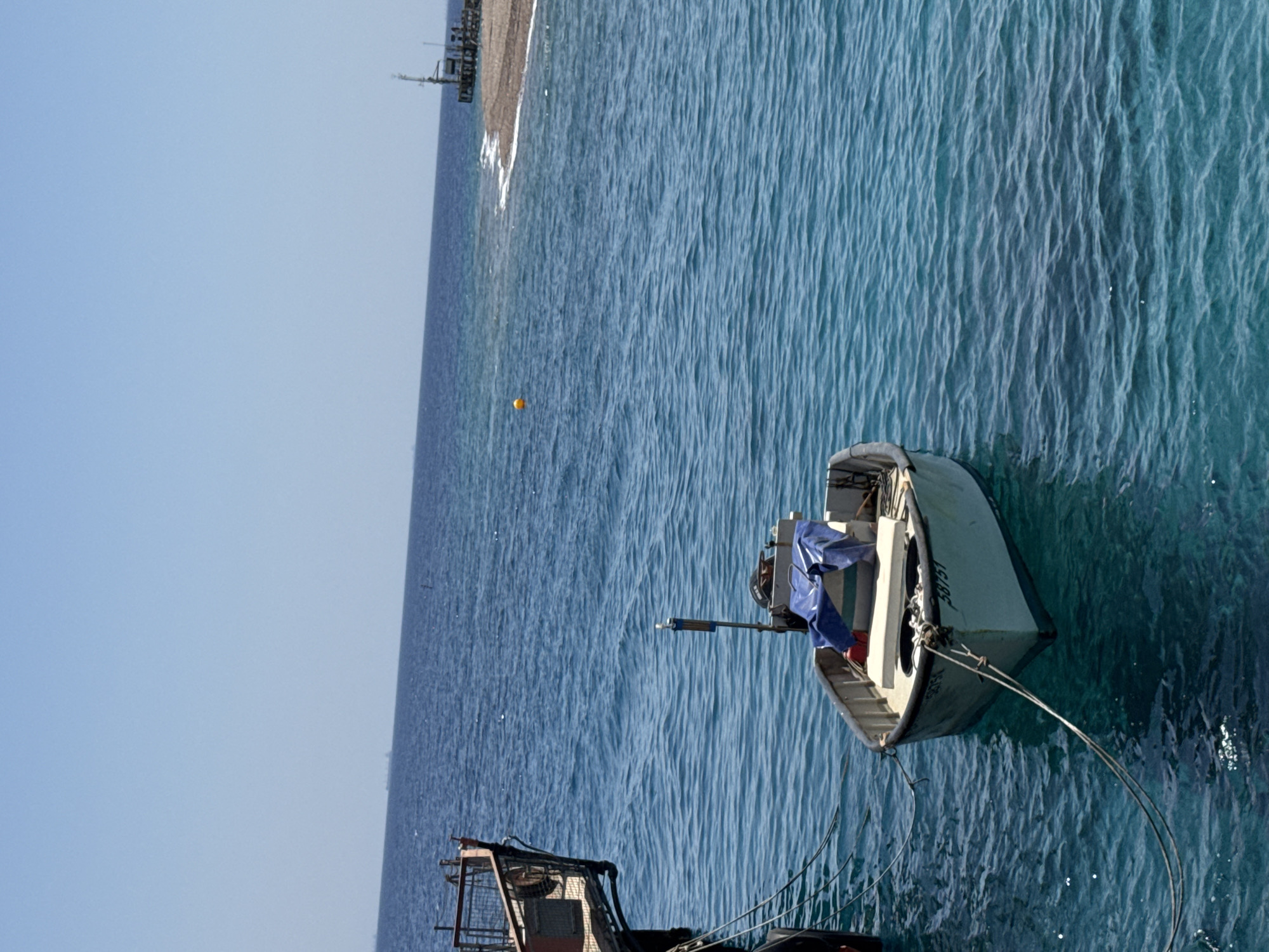 The image depicts a serene marine scene with a small white boat anchored in clear blue water. The boat has a blue tarp covering some of its contents and is tied to a larger docked structure on the left. In the background, a distant landmass with a pier and a navigational aid is visible, suggesting a coastal area. The sky is a pale, cloudless blue. The water exhibits ripples, indicating a calm day. The boat has the number "58751" visible on its side. The overall atmosphere is tranquil and suggests a port or harbor environment in Eilat, Israel, a city known for its Red Sea coastline.