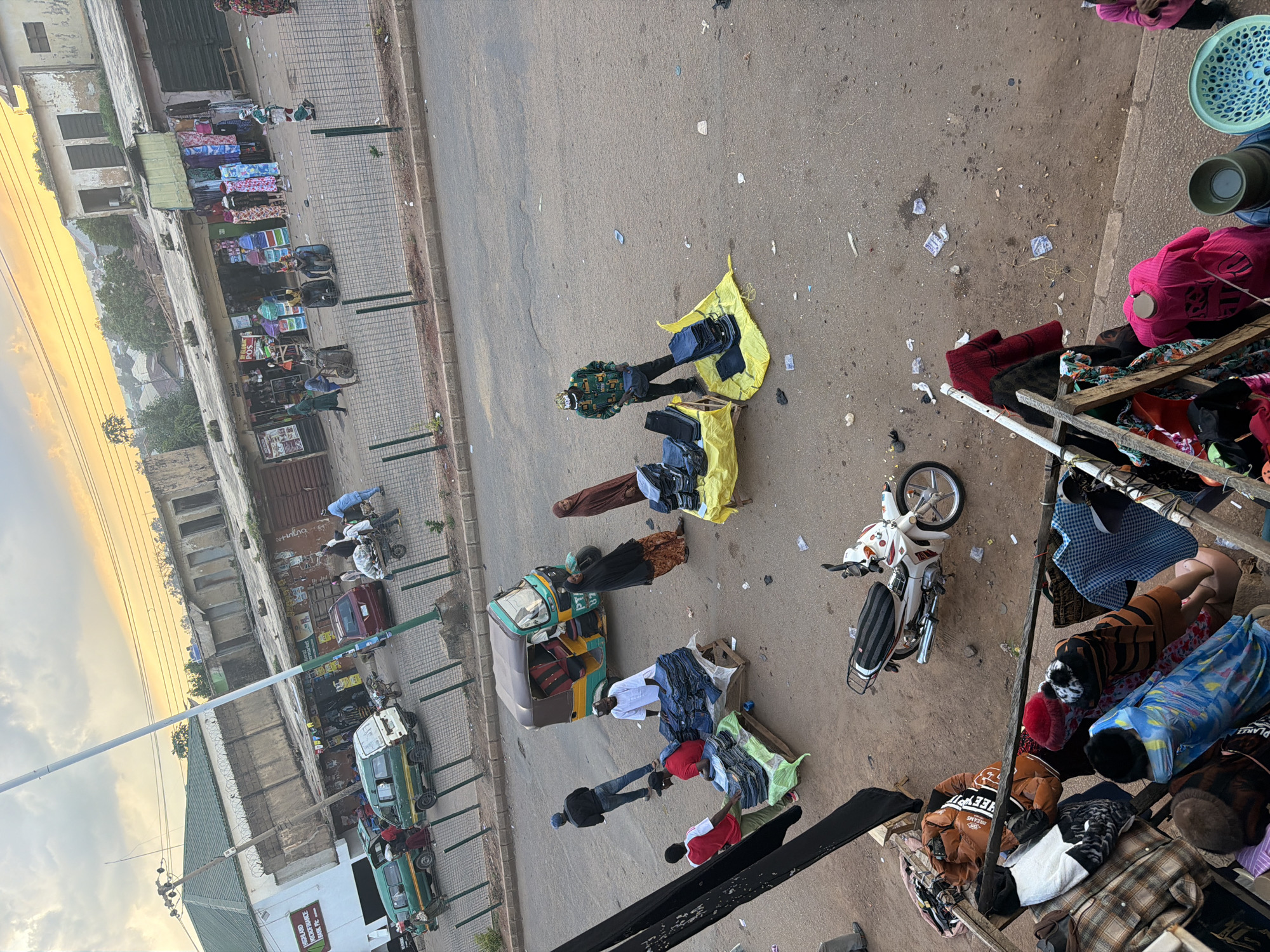 This is an outdoor, slightly elevated shot of a bustling street scene in Bukuru, Nigeria. The image is oriented vertically. The foreground features a wooden stall overflowing with various garments, including jackets, shirts, and blankets, suggesting a market or roadside vendor. To the right, a blue plastic basket and a dark green funnel-like object are visible.
In the middle ground, several individuals are engaged in commerce. A person in a patterned shirt stands next to a yellow tarp spread on the ground, displaying folded items, possibly clothing or textiles. Nearby, a tricycle, commonly known as a "keke," is loaded with luggage. Another person is seen tending to goods laid out on a yellow sheet. A white scooter is parked on the asphalt.
Further back, a row of shops or market stalls lines the sidewalk, displaying more goods, including what appears to be mattresses or large fabric rolls. Several vehicles, including green minibuses and a red car, are parked along the roadside. The backdrop includes buildings with weathered facades and a wide expanse of sky bathed in the warm, golden light of either sunrise or sunset. The overall atmosphere is one of vibrant activity, characteristic of a busy market day in a Nigerian town. The presence of numerous people and merchandise implies ongoing trade and daily life.