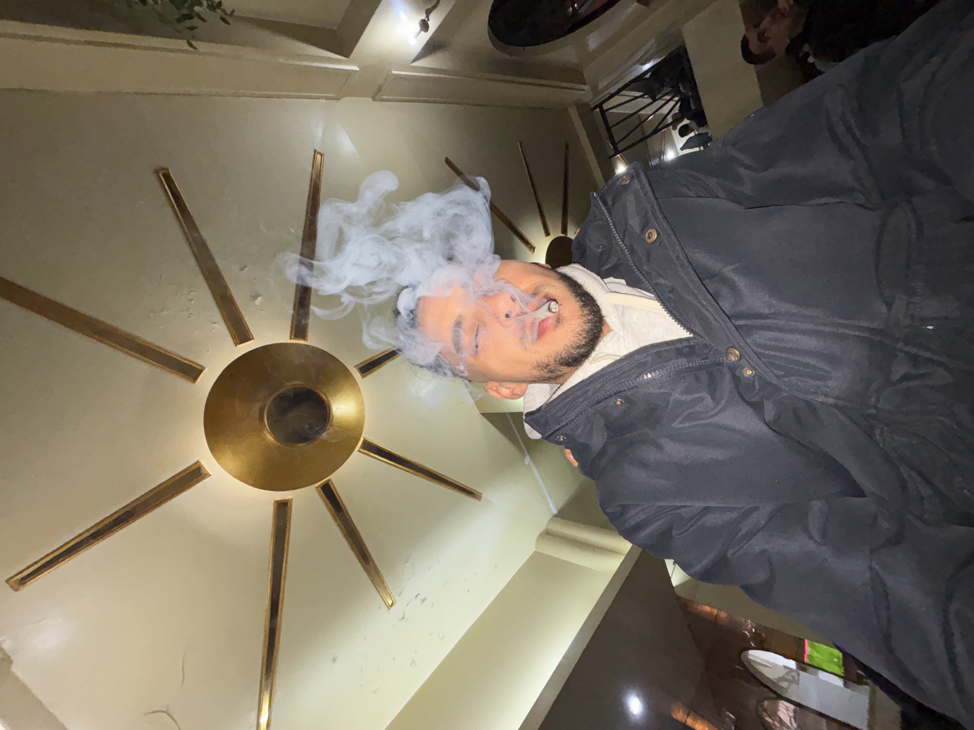 A low-angle, wide-angle indoor shot captures a male individual in the central foreground, exhaling a dense plume of white smoke from a cigarette held between his lips. He has a beard and short dark hair, wearing a dark puffer jacket over a light-colored hooded sweatshirt, with his eyes partially closed. Above him, a distinctive brass-colored ceiling fixture, resembling a sunburst or star with a central circular hub and radiating rectangular arms, is mounted on a light-colored ceiling. The background reveals a multi-level interior space, likely a cafe, restaurant, or lounge located in Tanger, Morocco. Visible elements include a dark-railed staircase leading to an upper seating area with blurred figures, light-colored architectural pillars, and subtle ambient lighting. The specific name or address of the establishment within Tanger is not discernible from the image.