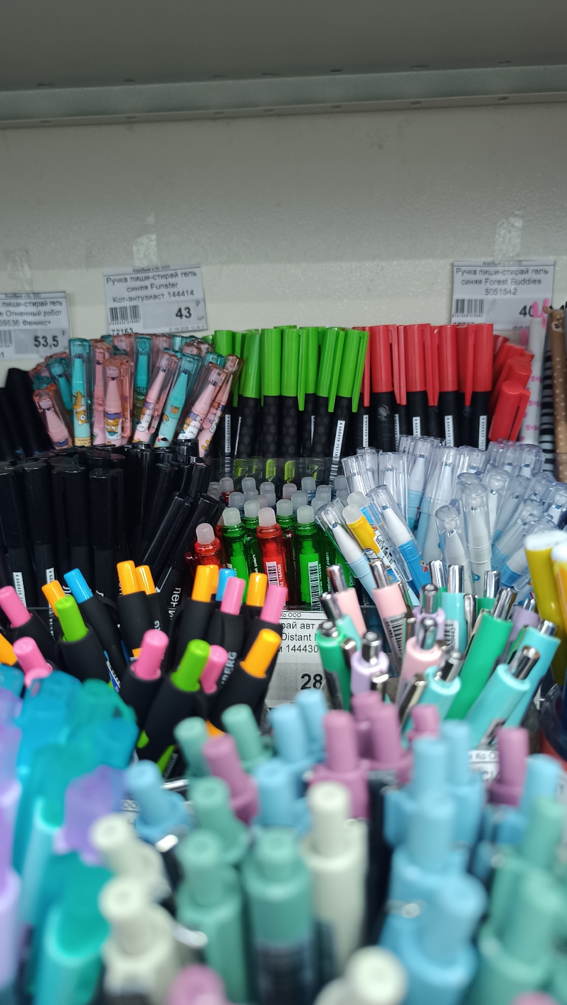 The image shows a shelf in a store with numerous colorful pens. There are multiple rows of pens with different colored caps and bodies, including pink, green, red, blue, and yellow. Some pens are in clear plastic packaging, while others are displayed individually in containers. Labels with prices and product information are visible on some of the pens. The background is a plain white wall. This scene appears to be a retail display of writing instruments in a store located in Chaikovsky, Russia.