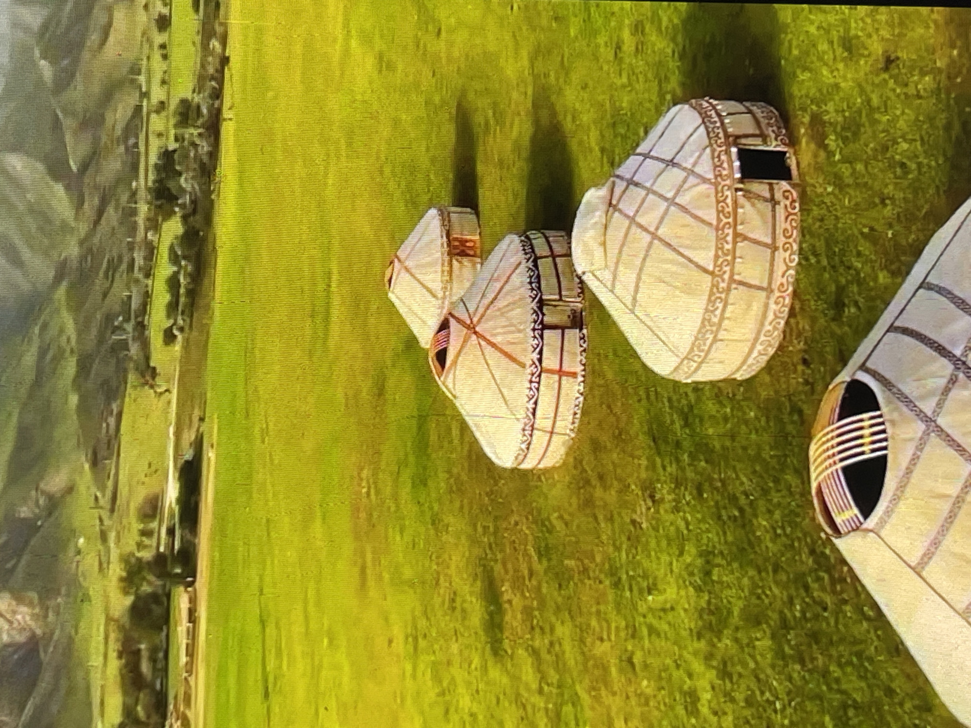 The image depicts a picturesque, mountainous landscape in Kyrgyzstan, featuring several traditional yurts scattered across a vibrant green field. In the foreground, a portion of a yurt is visible, its lattice structure and decorative patterns hinted at. Further back, three more yurts are arranged in a line, their conical roofs and patterned exteriors clearly defined against the lush grass. The yurts exhibit intricate detailing, including decorative borders and openings that suggest entrances. Beyond the yurts, the terrain slopes upwards, revealing a verdant valley dotted with trees and sparse vegetation. In the distance, imposing mountains with undulating slopes dominate the horizon. The overall scene evokes a sense of rural tranquility and cultural heritage, likely representing a nomadic or semi-nomadic dwelling in the Kyrgyz countryside. While the image captures a typical scene from Kyrgyzstan, it does not specifically indicate the location within Bishkek City, as yurts are more commonly associated with rural and nomadic lifestyles outside of urban centers.