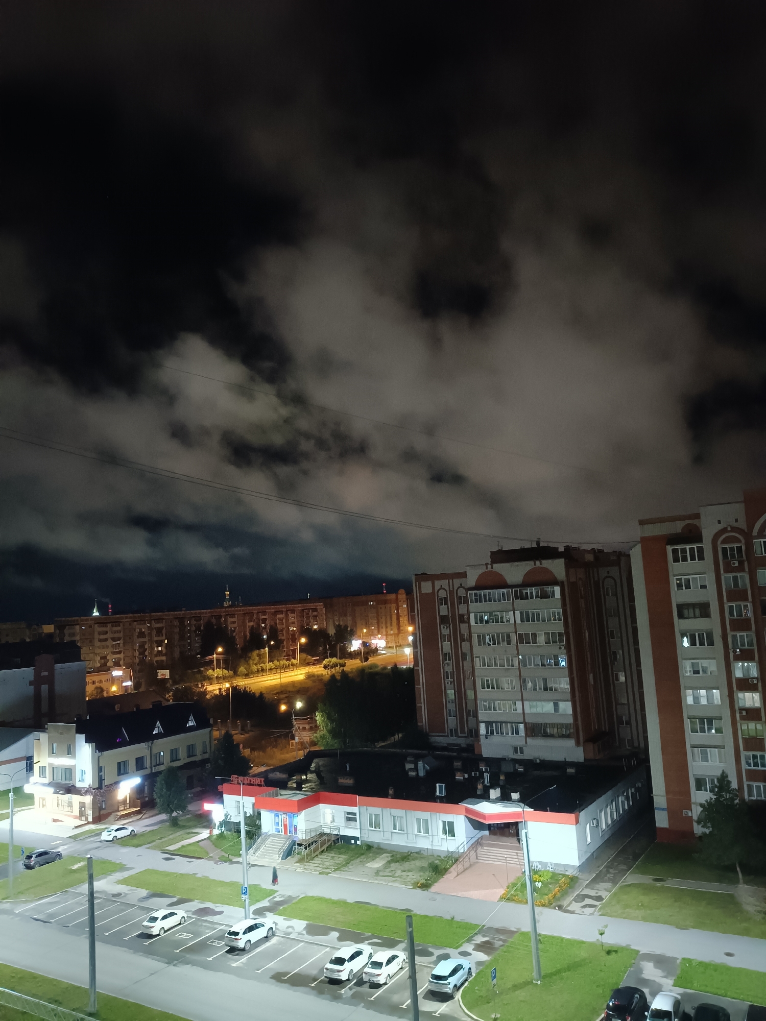 This is a nighttime, eye-level, full shot of a residential area in Yoshkar-Ola, Russia. The sky is dark with illuminated clouds. In the foreground, there is a parking lot with several cars parked. To the right, there is a commercial building with a red and white facade and illuminated windows. Behind it, there is a tall, brick residential building. In the background, there are more residential buildings and streetlights. The overall scene is illuminated by artificial lights, creating a contrast between light and shadow.