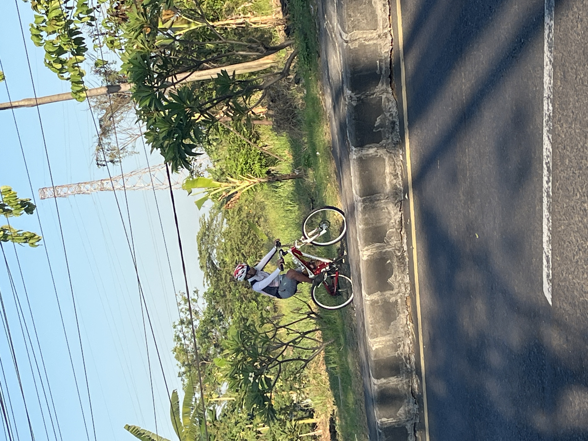A person wearing a helmet and athletic clothing is riding a red and white bicycle on a paved path next to a road. The person is on the left side of the bicycle, pedaling forward. The bicycle has two wheels, handlebars, and a frame. The path is bordered by a curb and then a grassy area with trees and bushes. In the background, there is a tall metal tower and power lines against a clear blue sky. The scene appears to be in a semi-rural or suburban area, possibly Siring, Indonesia, given the vegetation and the presence of a utility tower. The road in the foreground is asphalt with yellow and white markings. Sunlight casts shadows across the road, indicating it is daytime.