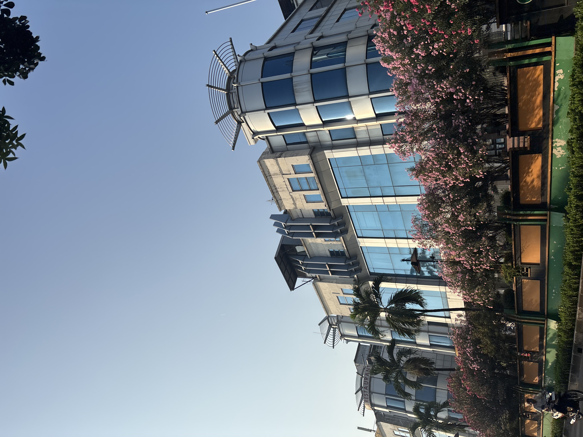 This is a low-angle, medium shot of a modern building in West Jakarta, Indonesia, under a clear blue sky. The building is a multi-story structure with a facade of blue glass windows and light-colored concrete or metal panels. The building has several architectural elements, including rounded corners and a circular structure with a metal frame on the rooftop of one section. Palm trees are visible in front of the building, and a line of blooming pink trees lines the foreground. A person is riding a motorcycle on a road in the lower left corner of the image, moving from left to right. A green median with decorative panels separates the road from the building and landscaping. The overall impression is one of an urban environment with commercial buildings and greenery.