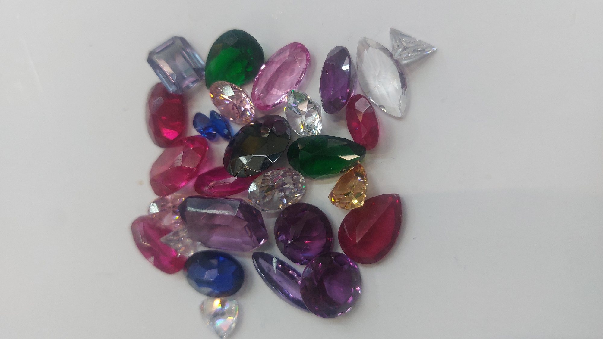 The image depicts a collection of loose gemstones scattered on a white surface. The gemstones vary in color, including shades of green, red, pink, purple, blue, yellow, and clear. The gemstones have different shapes, such as oval, pear, square, and round. No people, actions, or interactions are discernible in the image. There is no indication of a location in Novosibirsk, Russia or any other specific location.
