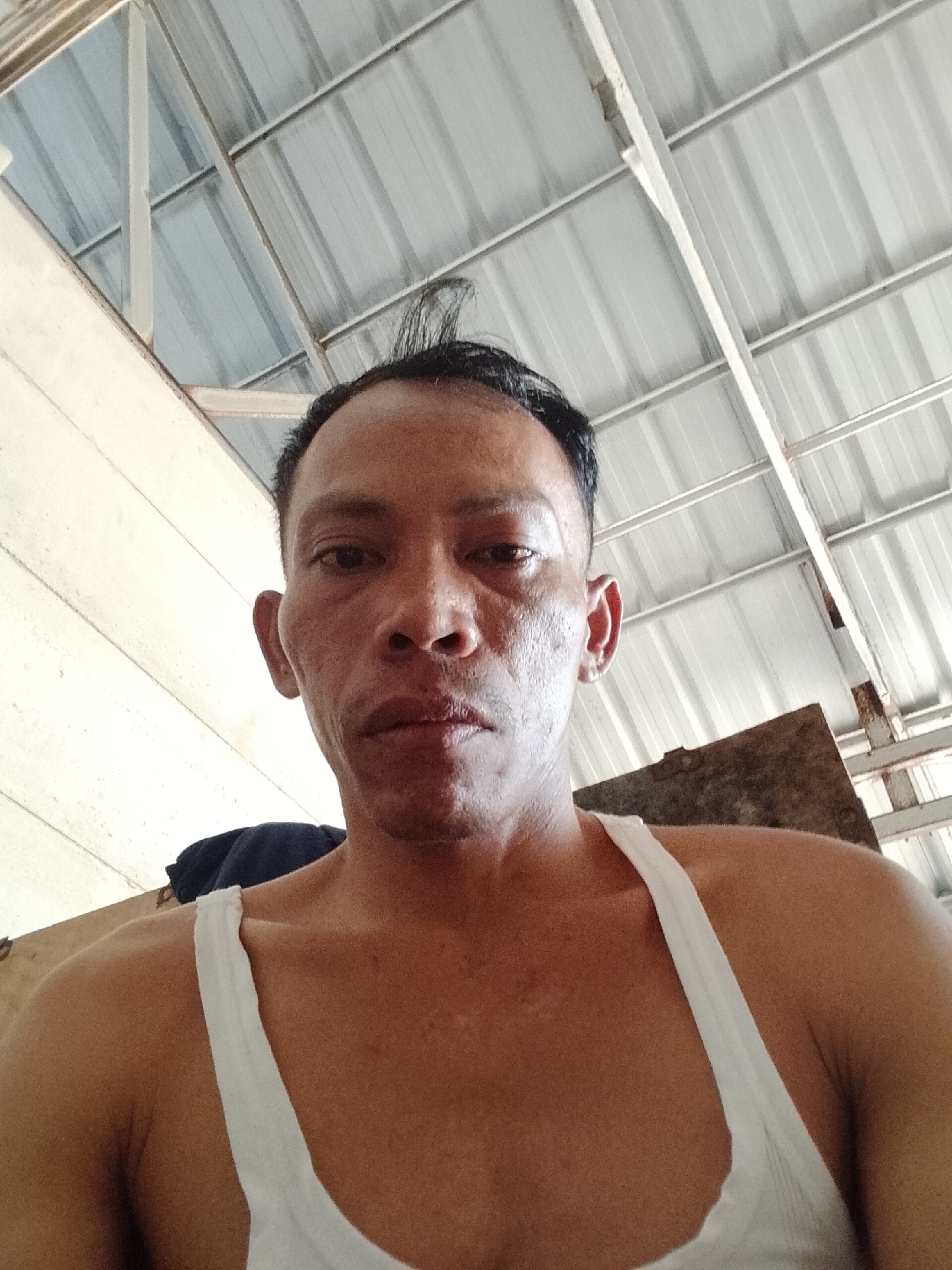 An extreme close-up, waist-up, selfie-style photograph captures a man from Bukit Ketok, Indonesia. He is wearing a white tank top, the straps of which are visible on his shoulders. His skin appears tanned and textured. He has short, dark hair, with a few strands sticking up on the crown of his head. His facial features are visible, including his eyes, nose, and mouth. The background of the image shows a corrugated metal roof supported by white beams. To the left, a textured wall is visible. The lighting suggests an indoor setting, possibly a workshop or storage area.