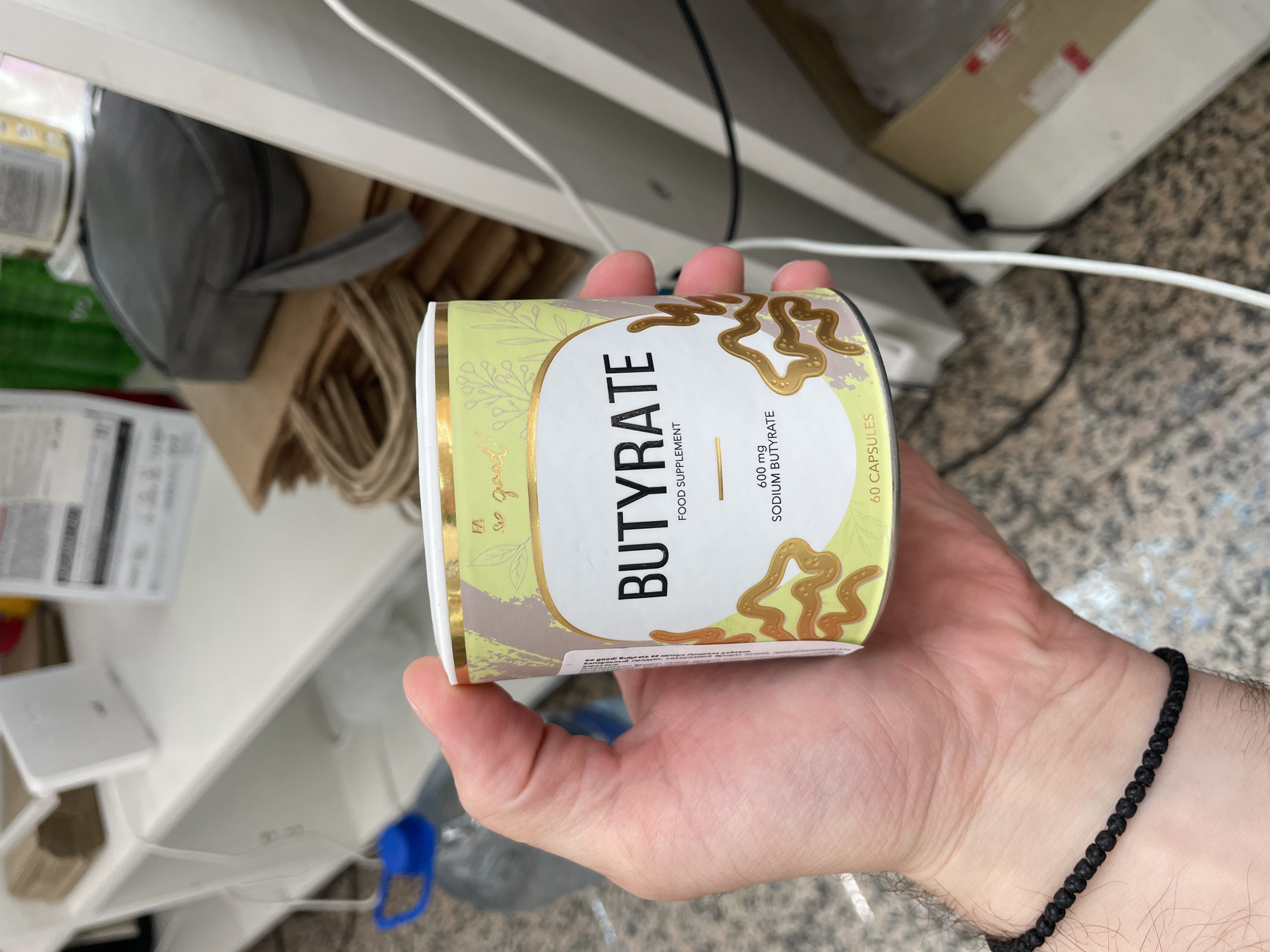 A person's hand, wearing a black beaded bracelet on the wrist, holds a cylindrical container of "BUTYRATE" food supplement. The container has a light green and brown abstract design with gold accents and text. The text on the container reads "BUTYRATE," "FOOD SUPPLEMENT," "600 mg SODIUM BUTYRATE," and "60 CAPSULES." The background suggests an indoor environment with shelves, stacked paper bags, a water bottle with a blue cap, and electrical cords. The image is taken from a close-up perspective, focusing on the hand and the product.