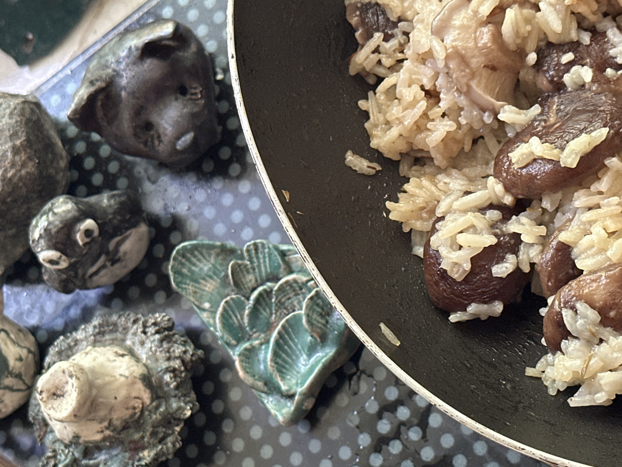 The image shows a partially filled frying pan of rice and mushrooms, sitting on a surface patterned with dark gray polka dots over a bluish-gray background. Next to the frying pan are several small, pottery figures. The figures are of various shapes, including an animal head, a round figure with prominent eyes, and a green, scale-like shape. The location is ตำบลเกาะพะงัน, Thailand.