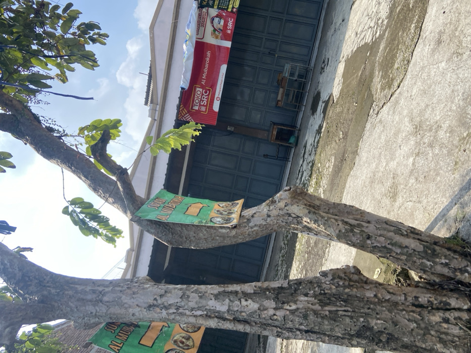 The image shows a tree trunk in the foreground, with a concrete driveway and a building in the background. The tree trunk is large and brown, with rough bark. It is split into two main branches. The driveway is cracked and gray, with a few patches of green moss. The building is a single story, with gray siding and several windows. There are two signs on the building, one for a store called "Toko SRC Al Mubarakah," and another for a store called "Sumber Berkah Mart." The signs are both red and white. There is no one present in the image. The location is a residential area in the city of Margaasih, Indonesia, with coordinates -6.942 latitude and 107.551 longitude.
