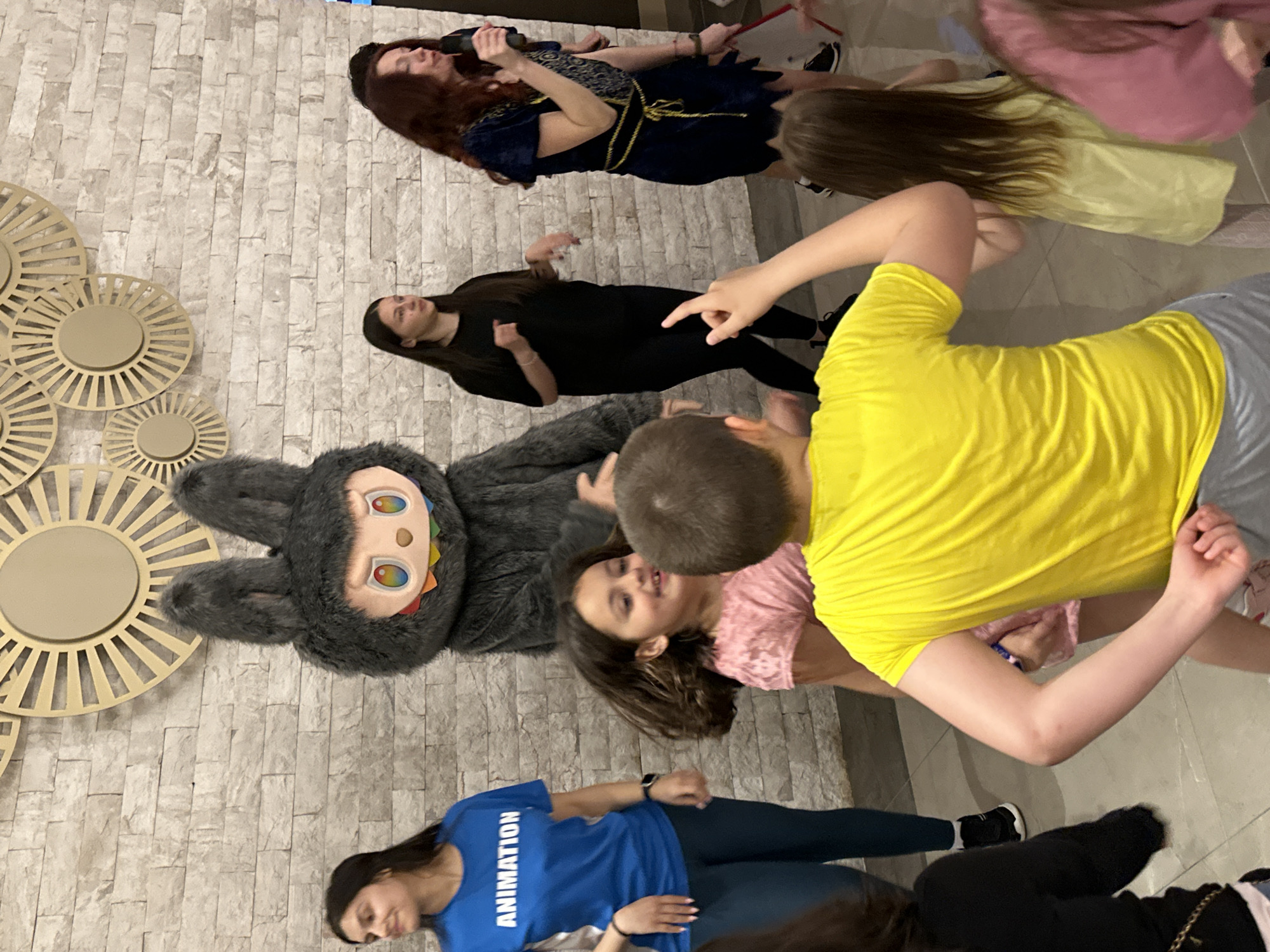 Scene in Kazanlak, Bulgaria:
A group of people are on the floor in what appears to be an indoor location with light-colored brick or stone-like wall with round, decorative, metallic sunburst accents.
People:
- Several individuals are lying on the floor.
- One woman wears a blue T-shirt with the word "ANIMATION" printed on it.
- Another woman is wearing a navy blue dress with gold accents and holding a microphone or similar object.
- A person wears a yellow shirt.
- Some people are partially obscured by other individuals or the camera angle.
Subjects:
- A large, gray, plush rabbit-like character with a colorful facial design lies on the floor.
Objects:
- Decorative metallic wall art with a sunburst design.
- Clothing worn by the individuals present.
Actions:
- People are lying down or kneeling on the floor, with some appearing to be interacting with each other.
- The woman in the blue dress seems to be speaking into the microphone.
Interactions:
- People are in close proximity to one another.
- There is apparent interaction between people and the costume.
Environment:
- Indoor area with a light-colored brick-like wall and some lighting fixtures.
- The floor surface is a light color.