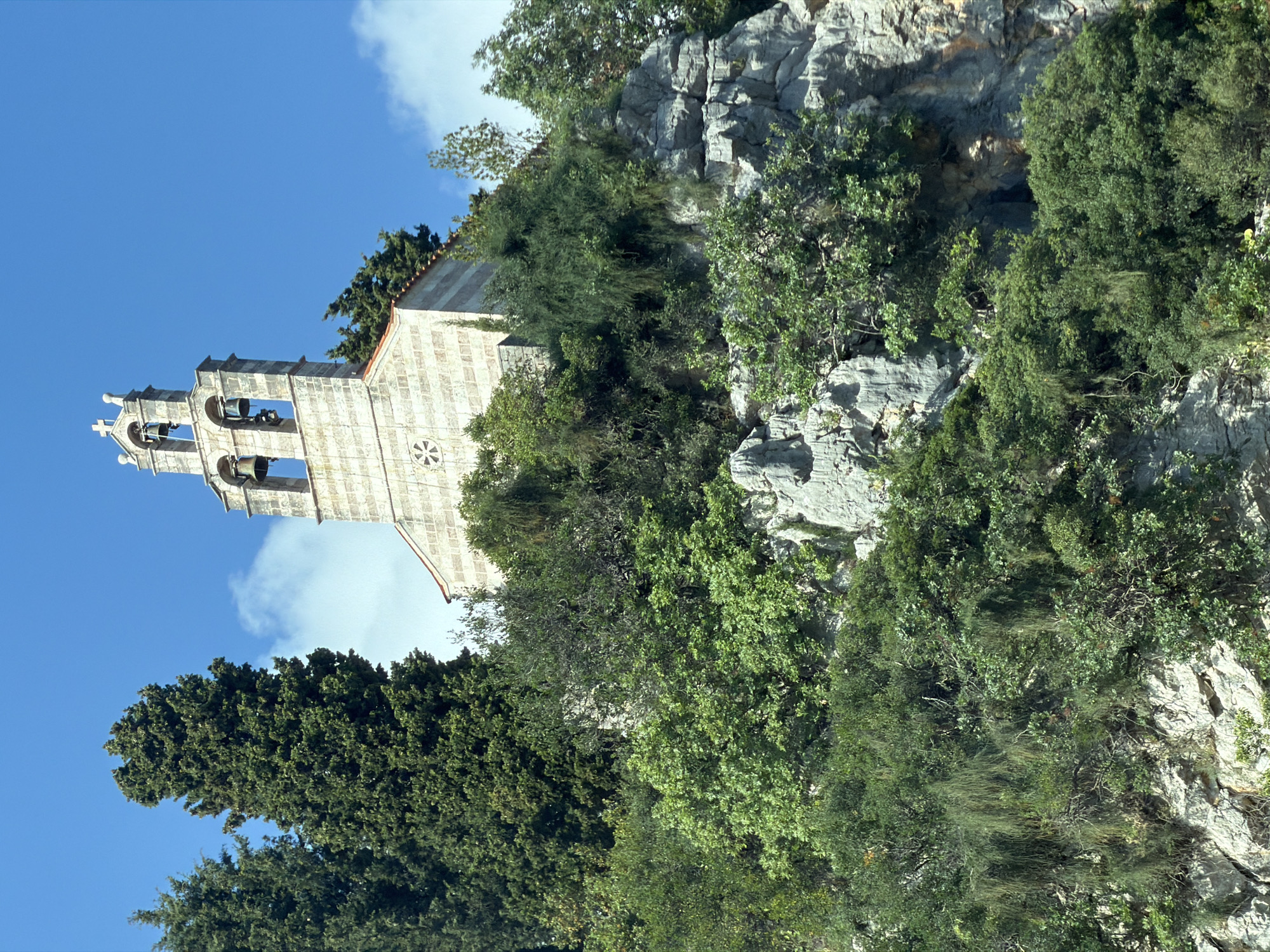 An image, correctly oriented, captures a stone church nestled on a rugged, tree-covered hillside under a bright blue sky with scattered white clouds. The setting is Kaluđerac, Montenegro, known for its scenic coastal and mountainous landscapes.
The central subject is a traditional church, constructed from light-colored stone blocks. Its most prominent feature is a tall, ornate bell tower (campanile) rising from its left side, composed of three visible levels of arched openings, each housing one or more dark bells. The church roof is terracotta-tiled. A circular decorative element, possibly a rosette window, is visible on the main facade of the church building.
The church is surrounded by dense, varied green vegetation. Taller, darker coniferous trees, likely cypresses, are visible on the lower left foreground, while a mix of lighter green bushes and smaller trees cling to the rocky terrain. Jagged, gray rock formations and cliffs form the backdrop and integrate with the vegetation around the church, suggesting it is built directly into the natural slope.
The time of day appears to be bright daytime, likely late morning or early afternoon, with clear weather and ample sunlight casting subtle shadows. There is no human activity or visible text in the image. The scene conveys a sense of tranquility and the harmonious integration of ancient architecture within a wild, natural environment.