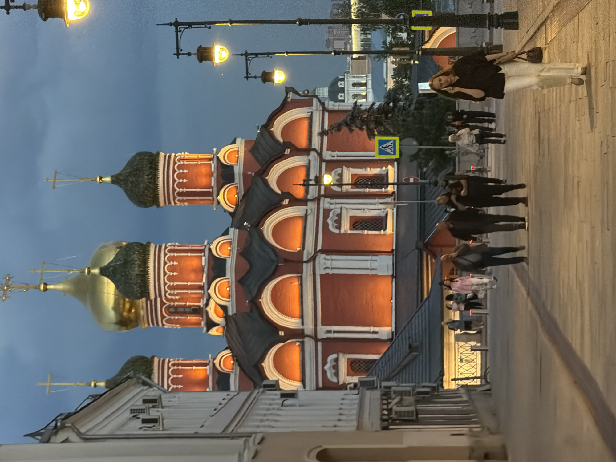 The image captures an evening scene in Moscow, Russia, featuring a prominent red and white church or cathedral at dusk. The sky is a deep blue, indicating either late evening or early night. Artificial lighting from streetlamps and internal illumination highlights the architectural details of the buildings.
The central subject is a large, multi-domed church with vibrant red walls and ornate white decorative trim. It features at least three distinct onion domes; the two larger ones are golden, reflecting the light, while a smaller dome on the far left is dark green. Each dome is topped with a golden cross. The church's facade is intricately designed with multiple arched windows and decorative elements, glowing warmly under the artificial light.
On the right side of the image, a paved sidewalk stretches into the distance, occupied by several pedestrians. In the mid-ground, a woman with auburn hair, dressed in a dark top and light-colored pants, stands facing the camera while holding an object to her ear, possibly a phone. She carries a dark handbag over her shoulder. Further down the sidewalk, other groups of people are seen walking.
Illuminated black lampposts with bright yellow lights line the street. A blue square sign with a white pedestrian crossing symbol is visible on a pole near the sidewalk. In the background, beyond the church and possibly across the Moskva River, distant buildings and lights are faintly visible, along with some trees. In the foreground on the left, the roof and upper part of another building with a gray roof are partially visible.