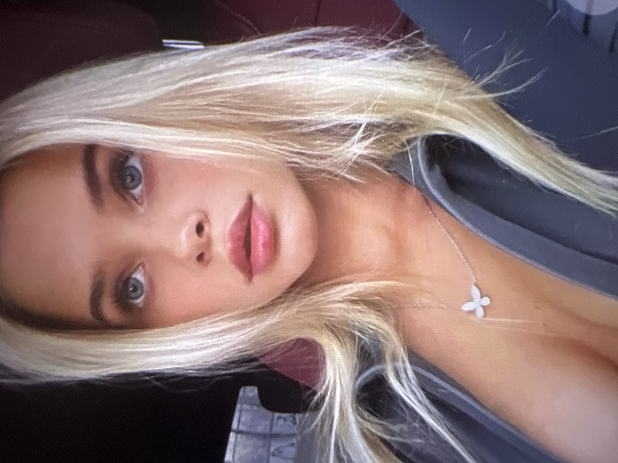 The image shows a close-up of a young woman with blonde hair and blue eyes, looking upwards. She is wearing a grey jacket and a delicate silver necklace with a butterfly pendant. Her makeup is subtle, with emphasis on her lips and eyes. The background is dark and blurred, with a hint of red upholstery suggesting she might be in a car. There is no information or visual evidence in the image that places it in Pyatigorsk, Russia, or any other specific city.