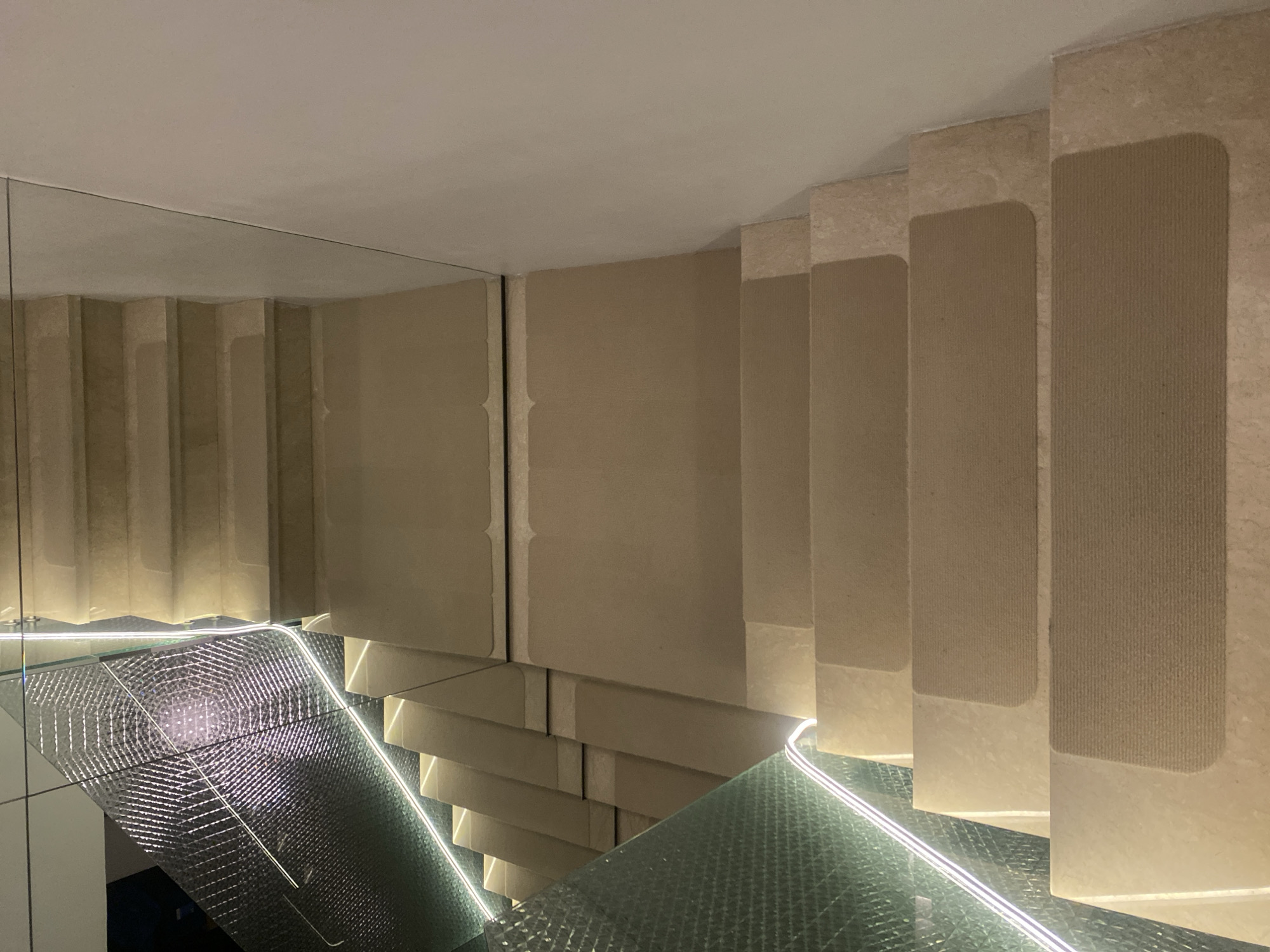 This image shows a modern staircase with marble steps and a glass railing. The staircase is illuminated by integrated LED lights, creating a sophisticated ambiance. The steps have non-slip treads for safety. The background features a textured, metallic paneling on the left and a plain white wall on the right, suggesting an interior setting. The perspective is looking down the stairs, emphasizing their width and the clean lines of the design. The overall impression is one of luxury and contemporary design, likely found in a high-end residential building, hotel, or commercial space in Kuala Lumpur, Malaysia. There are no people or animals present in the image, and no discernible actions are taking place.