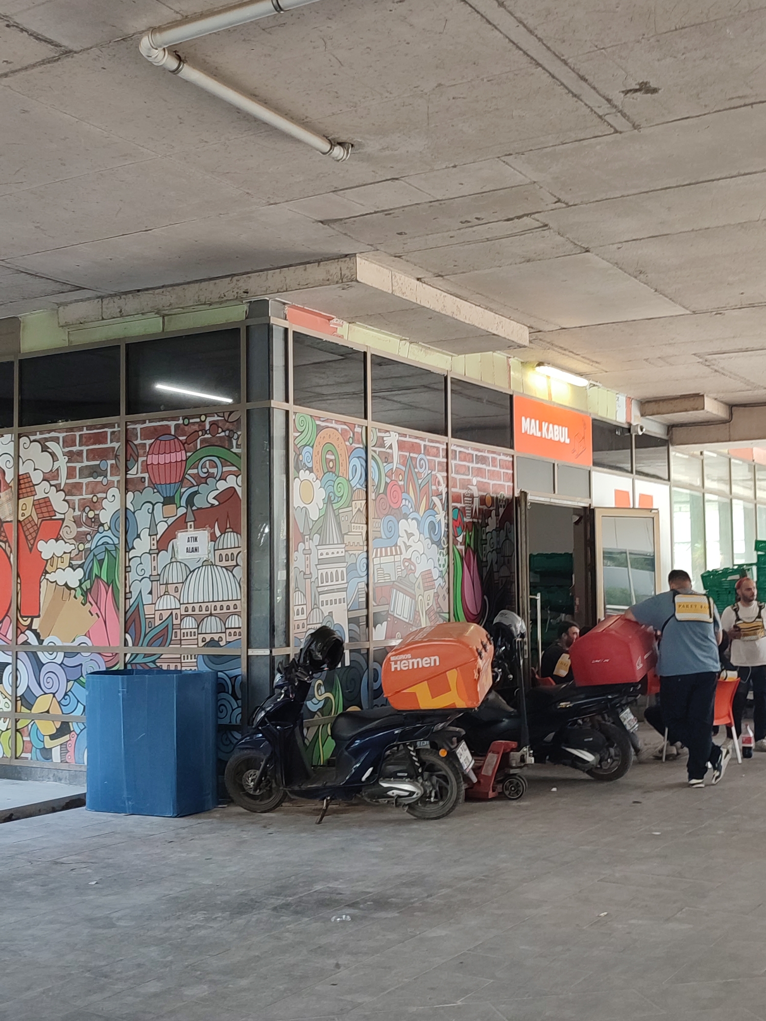 The image shows a street-level view of a building with large windows adorned with colorful murals depicting buildings, balloons, and abstract designs. Above the windows, an orange sign reads "MAL KABUL". Several parked motorcycles with delivery boxes are visible on the pavement in front of the building. Some people are standing and interacting near the entrance. The overall environment appears to be an urban setting, likely a commercial area in Kadıköy, Turkey, given the context of delivery services and the sign. The lighting suggests it's daytime.