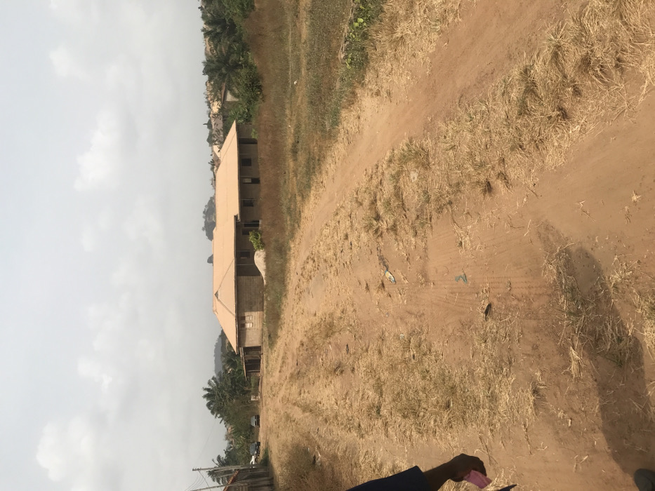 The image depicts an unpaved, reddish-brown dirt road in Akure, Nigeria, under a uniform overcast sky. The road exhibits distinct tire tracks and is interspersed with patches of dry, light-brown grass and sparse green vegetation. Scattered debris, including small pieces of plastic, is visible on the road surface. In the midground, a prominent single-story building constructed from grey concrete blocks features a distinct orange-brown corrugated metal roof. This structure appears to be under construction, with open window frames and no glass installed, and a visible pile of sand or aggregate positioned near its front. To the left of the main building, further down the road, several smaller structures and two parked vehicles are discernible. The background features undulating terrain with hills covered in dense green vegetation and additional distant buildings. In the immediate foreground, partially visible in the bottom left, is the lower right arm and hand of an individual wearing a dark blue sleeve, holding a small, rectangular pink object. No direct action or interaction is depicted by the person.