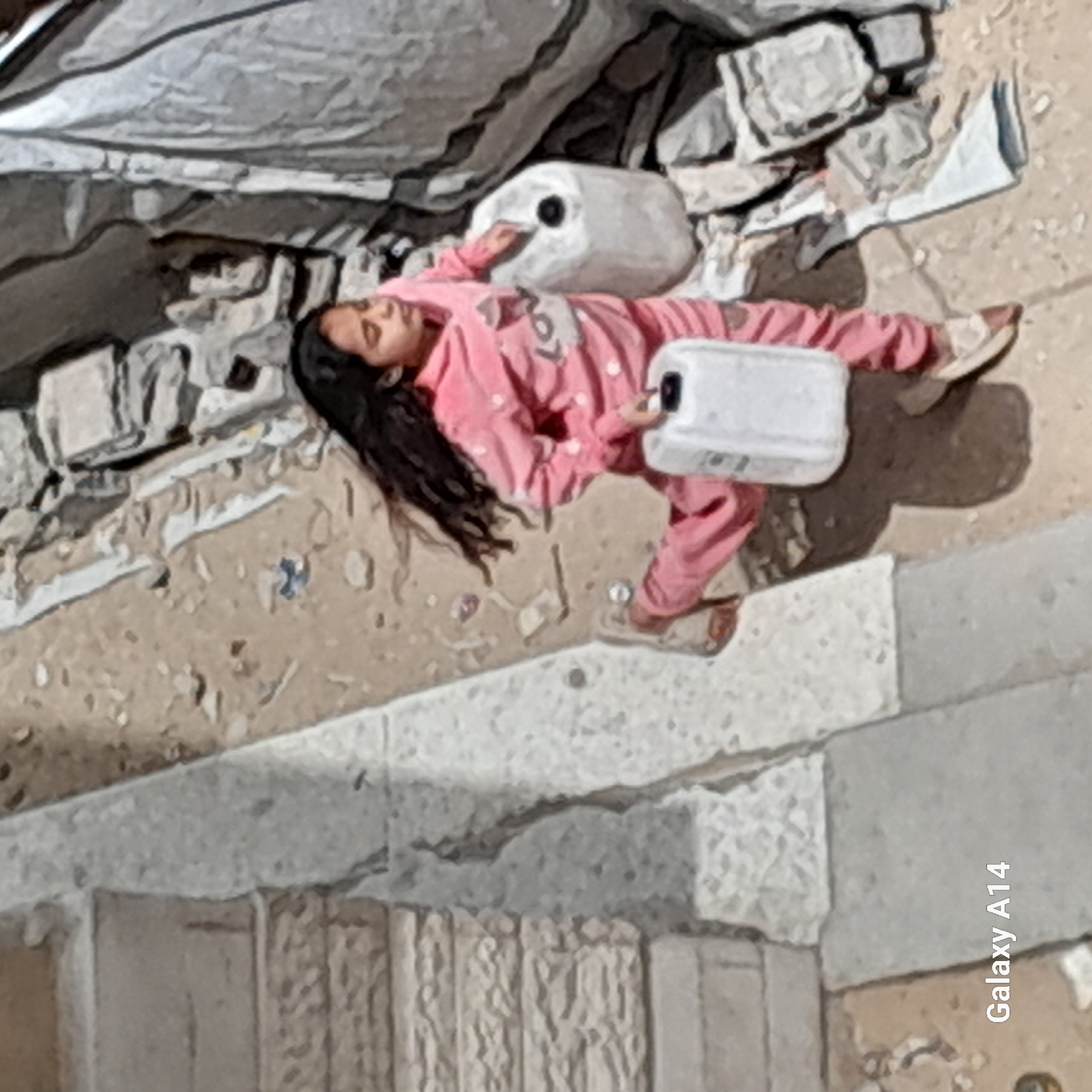 A young girl is depicted lying on her back amidst a scene of rubble and debris. She wears a pink long-sleeved top with white patterns and matching pink trousers. Her eyes are closed, suggesting she is sleeping or resting, and her dark hair spreads out around her head on the dusty ground. One white plastic jerry can or container is near her head, and another is held loosely by her right hand. The surrounding environment is characterized by broken concrete blocks, fragments of building material, and what appears to be a large, crumpled gray tarp or fabric in the background. The ground is uneven, composed of dirt and small stones. In the foreground and to the bottom right, light-colored concrete or stone steps/blocks are visible. The lighting indicates it is daytime. The overall scene evokes a sense of hardship and vulnerability. The text "Galaxy A14" is visible vertically along the right edge of the image.