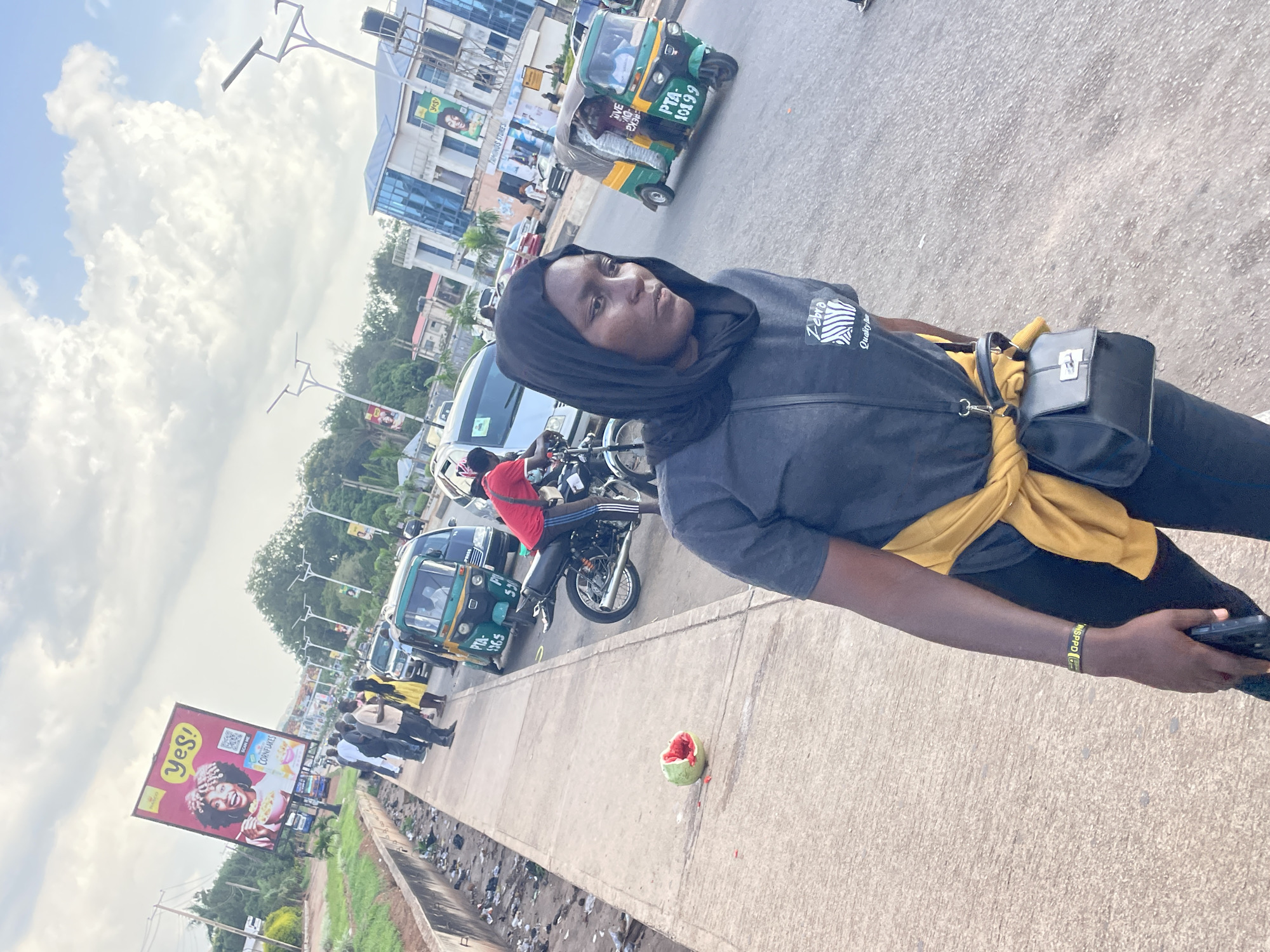 A young woman with dark hair and a black hijab is the main subject of the image, which appears to be taken outdoors in a busy urban environment, likely in Barakin Lerawi, Nigeria, given the context. She is wearing a dark grey t-shirt with a zebra-like graphic and the text "Quality Da" visible, and black pants. A yellow scarf is tied around her waist, and she has a black crossbody bag. She is looking towards the camera with a neutral expression.
The setting is a street with traffic. Several vehicles are visible, including a green and yellow tricycle (also known as an "okada" or "kekè") with license plate "PTA-10199", other tricycles, motorcycles, and cars. A billboard advertising "Yes! Cornflakes" is prominently displayed on the left side of the image, featuring a woman with curly hair holding a bowl of cereal. There are also buildings and trees in the background, suggesting a commercial or residential area. The sky is partly cloudy, indicating it might be daytime. The ground is paved, and there is some litter visible along the side of the road. A partially eaten piece of fruit, possibly a watermelon, lies on the pavement near the woman. The overall scene suggests a typical day in a bustling Nigerian town.