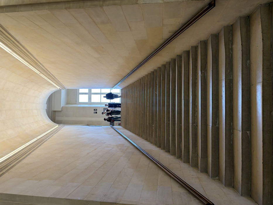 A long, wide stone staircase ascends under a high, vaulted stone ceiling within the Louvre Museum, Paris, France. The staircase consists of approximately twenty-six visible light-colored, cut-stone steps, flanked by matching stone block walls. Dark wooden-topped metal handrails run along both walls, guiding the ascent. At the brightly illuminated landing at the top of the stairs, four individuals are present. From left to right, these include a person with dark hair and clothing carrying a light-colored shoulder bag, a person in a light top and dark trousers wearing a backpack, a person in dark clothing, and a larger person in dark outerwear. These individuals are either standing on the landing or commencing their descent. The landing is brightly lit by a tall, multi-paned window, which casts natural light down the upper section of the staircase. A red fire extinguisher on a stand is visible against the left wall at the top landing. The lower portion of the staircase is comparatively shadowed, while the upper steps are distinctly illuminated.