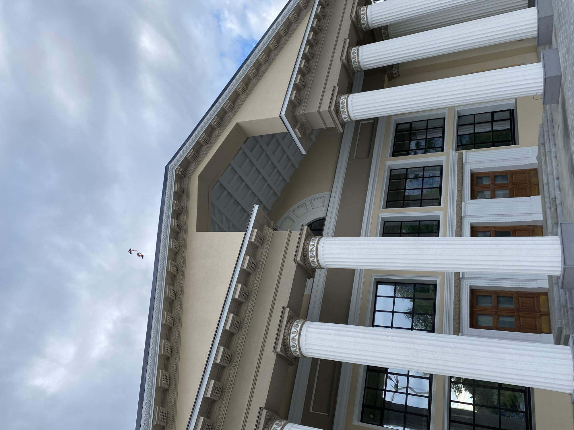 The image shows a low-angle, wide shot of the facade of the N.I. Lobachevsky Scientific Library in Kazan, Russia. The building is classical in style, with a beige exterior, large white fluted columns topped with ornate capitals, and a pedimented roof. Multiple tall, multi-paned windows with black frames are visible on the upper level, reflecting the cloudy sky and surrounding trees. Two wooden doors are situated at ground level between some of the columns. Two flags, one red and white, and another red, white and blue, are visible atop the roofline. The sky is overcast with some patches of blue. The scene is static, with no people or animals present.