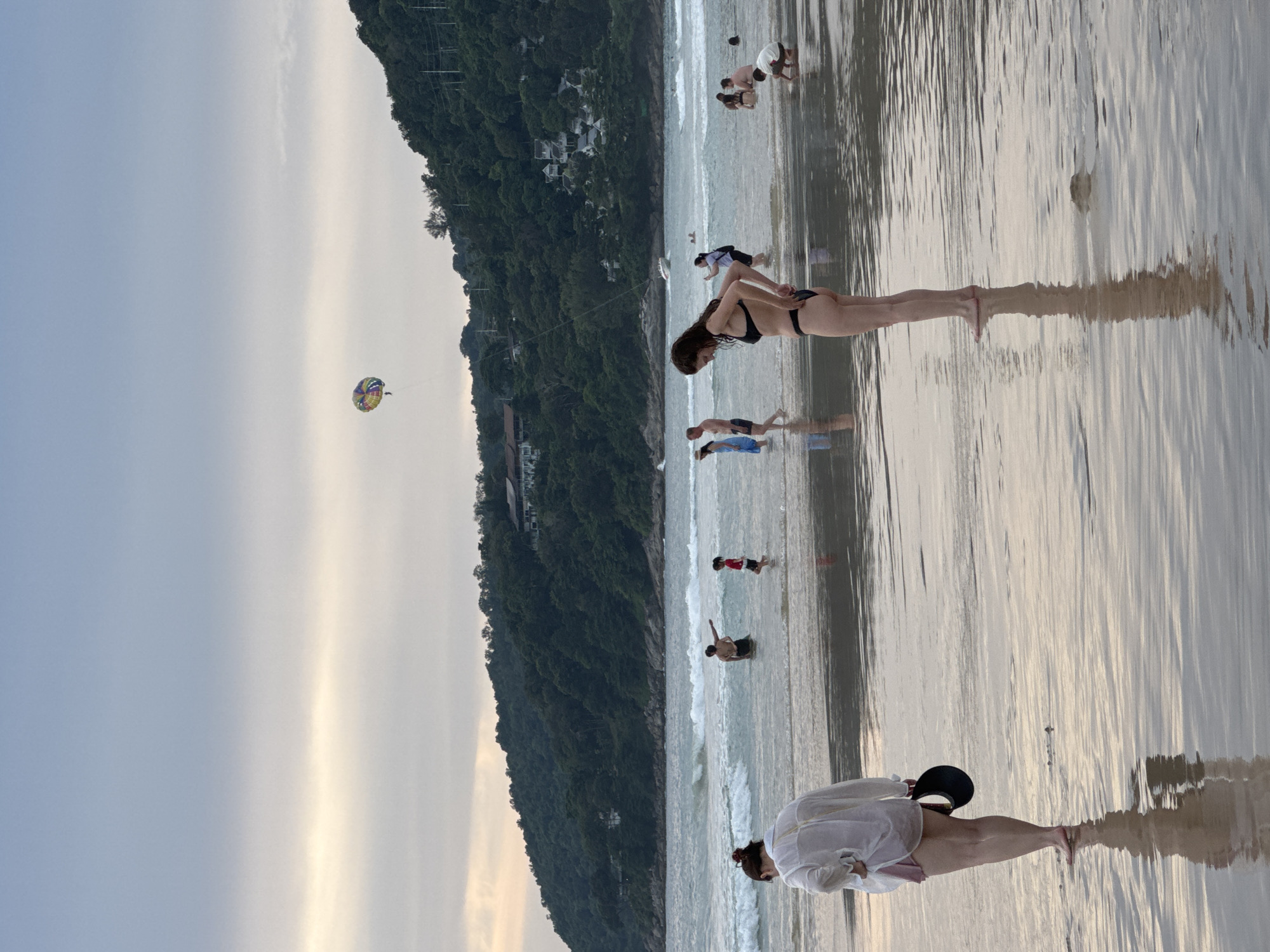 An aerial view of Karon Beach, Thailand, showcases a tranquil evening scene with a serene sky, calm waters, and a lush green hillside overlooking the shoreline. Several people are enjoying the beach and the ocean. A parasailer is visible in the sky, adding a splash of color and activity. The beach is wet, reflecting the soft twilight hues of the sky.