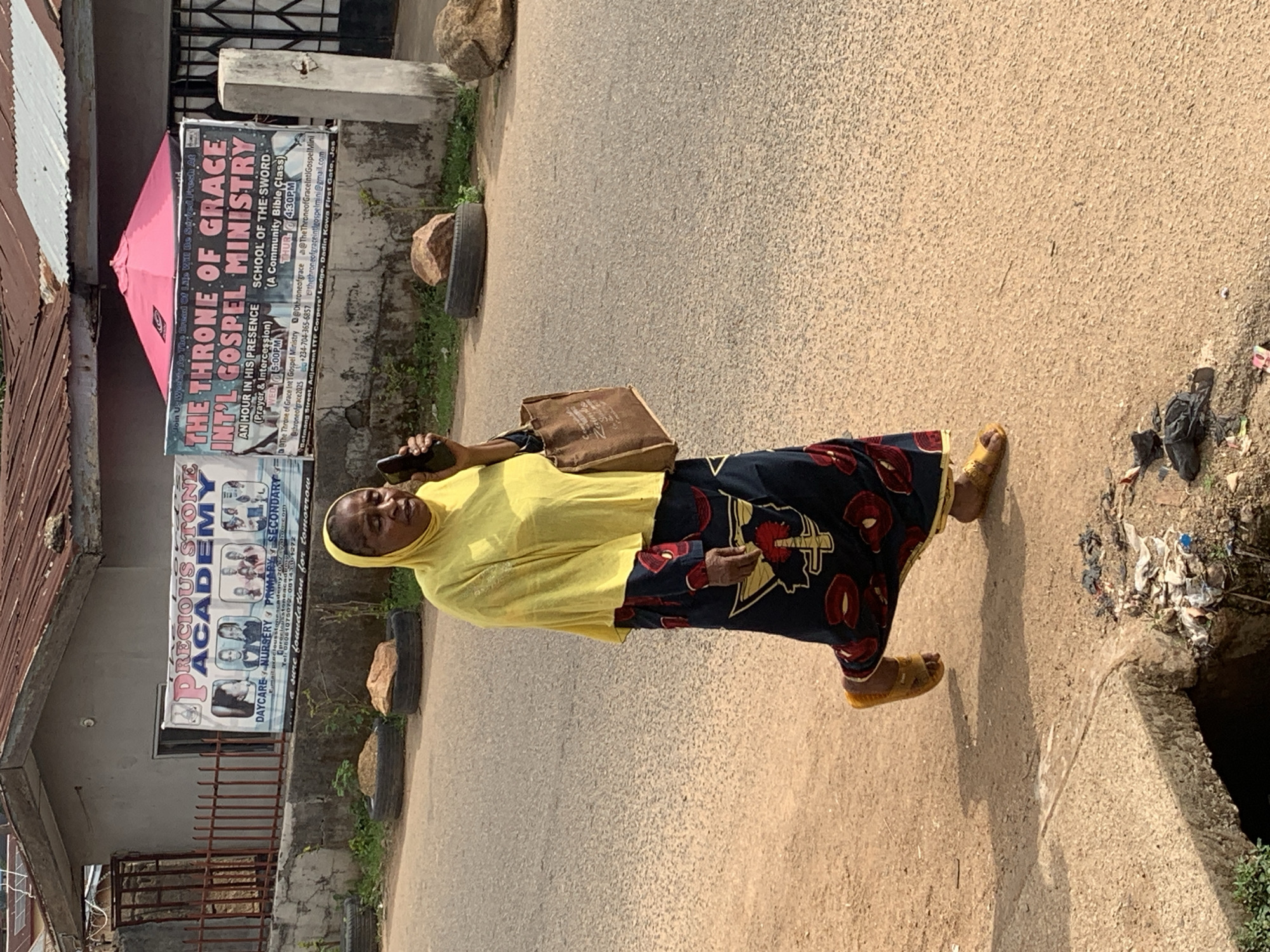 The image, oriented correctly, captures a bright, sunny daytime street scene in Jos, Nigeria. A woman, central to the composition, walks on a paved road. She is dressed in a yellow hijab, a long dark blue dress adorned with red circular patterns, and yellow sandals. She carries a brown shoulder bag and holds a smartphone in her right hand, looking towards the camera with a neutral expression. Her shadow is cast distinctly on the road.
On the left side of the road, behind the woman, are buildings with corrugated metal roofs. Two large banners are prominently displayed on these structures. The banner on the left advertises "PRECIOUS STONE ACADEMY," listing services such as "DAYCARE NURSERY PRIMARY SECONDARY" and promising "a sure foundation for tomorrow," along with phone numbers "08061075072" and "08143101272."
The banner on the right promotes "THE THRONE OF GRACE INT'L GOSPEL MINISTRY," inviting participation in "AN HOUR IN HIS PRESENCE (Prayer & Intercession) WED 5:00PM" and "SCHOOL OF THE SWORD (A Community Bible Class) THUR 4:30PM." This sign includes various contact details: social media handles (@Dthroneofgrace, @TheThroneofGraceIntiGospelMini, @throneofgrace2025), an email address (thethroneofgraceintlgospelmini@gmail.com), a phone number (+234-704-365-6857), and an address: "Botmang Street, Adjacent ITF Corpers' Lodge, Dadin Kowa First Gate, Jos."
Along the roadside, rocks and repurposed tires are visible, some possibly acting as makeshift planters or barriers. A small pile of litter lies near a drainage ditch on the right. The scene depicts a common street environment in a Nigerian urban or semi-urban area, characterized by local businesses and religious institutions.