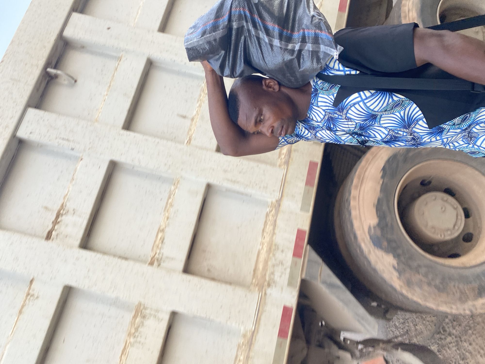A man stands next to a large truck in Abofifia, Nigeria. The man is carrying a grey plastic bag on his head. He is wearing a blue and white patterned shirt, black shorts, and a black satchel. The truck appears to be a dump truck, with a white bed marked with square patterns and red reflective tape. The truck's tires are dusty. The ground appears to be dirt or gravel.
