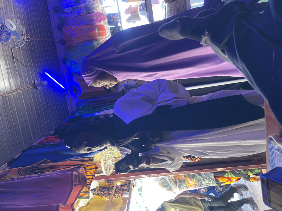 This image depicts an indoor market stall in Samaru, Nigeria, illuminated primarily by artificial blue light from an overhead LED strip. Two young females are prominent in the foreground. The female on the left, with dark skin, wears a black hijab, a white long-sleeved garment, and a black scarf. She is engrossed in a black smartphone held in both hands, with a red bracelet visible on her left wrist and a small light-colored bag hanging from her arm. The female on the right, also with dark skin, wears a light purple full-length hijab that covers her head and body; she is looking towards the right with an upward gaze, appearing to observe something beyond the frame.
The background features numerous colorful garments, including dresses and hijabs, displayed on racks and shelves, consistent with a clothing shop. A white ceiling fan with blue blades is mounted in the upper right. A dark wooden counter or surface is visible in the immediate bottom foreground, with the foot of a person wearing a dark sock and a decorative ring, suggesting the viewpoint of a seated individual. Through an open doorway or entrance on the far left, a male figure wearing a light green t-shirt, blue trousers, and sandals is partially visible, walking past. Another obscured figure in patterned clothing is behind him. The ceiling appears to be made of corrugated metal or a similar material.