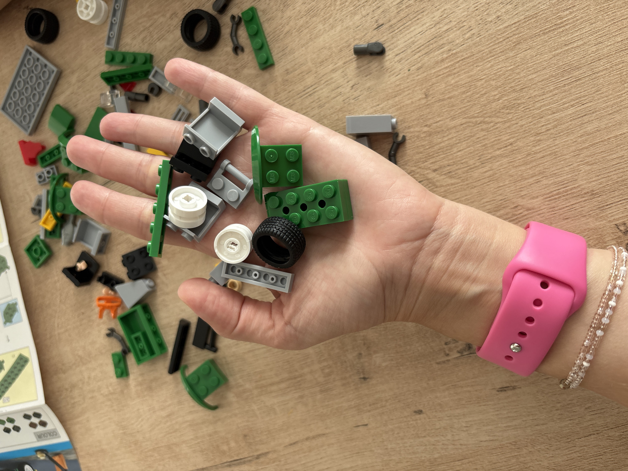 A person's hand is shown holding a pile of LEGO bricks over a wooden surface. The LEGOs include various shapes and colors such as green bricks, gray pieces, a black tire, and white circular pieces. A pink smartwatch is visible on the person's wrist. The background shows a wooden table with scattered LEGO pieces. The lighting suggests it is daytime.