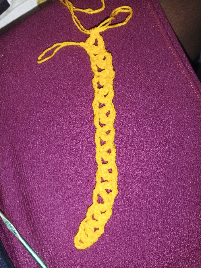 The image depicts a close-up view of a crafting scene, presumably located in Nsukka, Nigeria. A bright yellow, unfinished crocheted piece, approximately 20-25 cm in length, lies vertically on a textured deep purple fabric surface, slightly off-center to the right. The top end of the crocheted item shows loose yarn loops, indicating it is incomplete. A silver-colored metal crochet hook, bearing the inscription "4.0mm", is partially visible in the bottom-left corner, resting on the same purple surface. No individuals are present. The arrangement suggests a paused handcraft activity within an indoor environment.