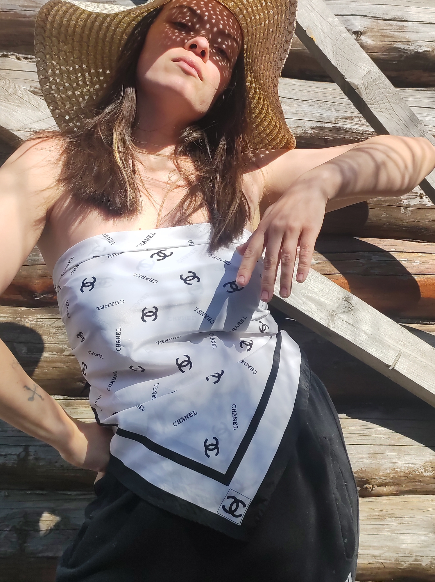 This media file captures a person posing outdoors in Фоки, Russia, on a bright, sunny day. The individual, appearing to be female, wears a wide-brimmed straw hat that casts distinct dappled shadows across her face and upper body. Her long, brown hair frames her face. She is dressed in a white and black scarf, stylized as a bandeau top, which prominently features the brand name "CHANEL" and its interlocking C's logo repeated across the fabric. Dark-colored bottoms are visible below the scarf. A tattoo depicting crossed bones is visible on her left forearm. She strikes a relaxed pose, leaning against a rustic wooden background composed of horizontal logs and some diagonal planks. Her right arm rests on one of these wooden beams, while her left hand is placed on her hip. The strong sunlight creates sharp shadows from both the hat and the wooden structure, highlighting the texture of the wood and the details of her attire. The visible text confirms "CHANEL" is displayed multiple times on her top.