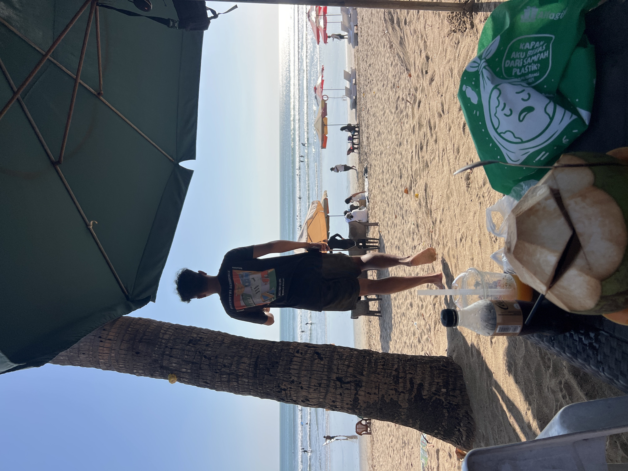 The image depicts a sunny beach scene in Kuta, Indonesia, with the ocean visible in the background. In the foreground, a table is set with a halved coconut, a bottle of Coca-Cola, a plastic cup with a straw, and a green tote bag with an Earth graphic and text that reads "AKU BISA TANPA PLASTIK?". A palm tree trunk is prominently featured on the left side of the frame, casting shadows on the sand.
In the mid-ground, a young man wearing a black t-shirt and shorts walks away from the camera towards the water. Several beach umbrellas and lounge chairs are scattered across the sand, with some people relaxing under them. The ocean is calm with gentle waves, and several individuals are seen in the water, some appearing to be surfers. The sky is clear and blue.