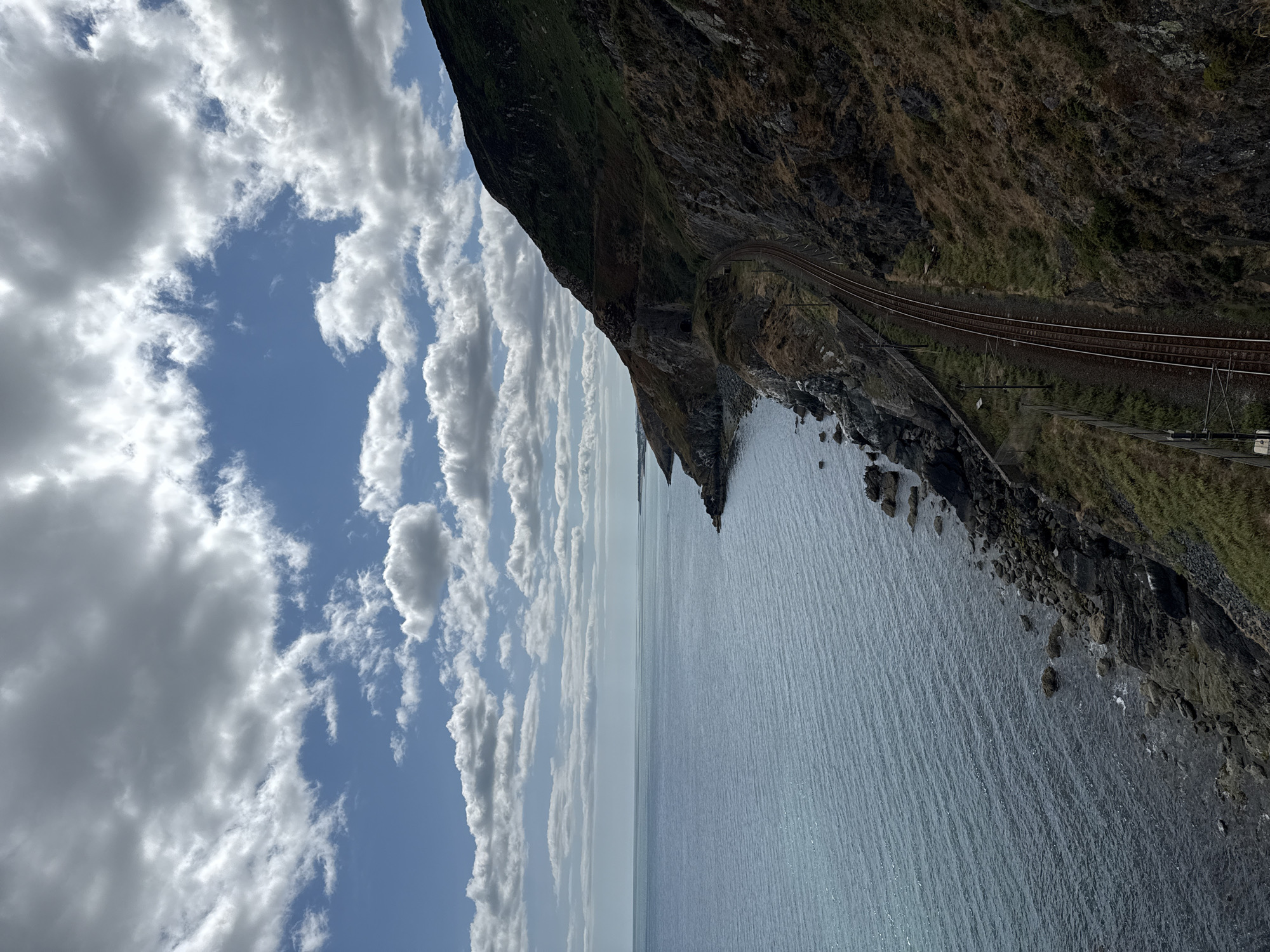 The image depicts a scenic coastal view along the railway line in Bray, Ireland. The camera is positioned high, looking down and to the right. The scene is dominated by the vast expanse of the sea on the left, with gentle ripples reflecting the sky's light. The coastline to the right is rugged and rocky, covered in sparse, dry vegetation. A railway track runs along the edge of the hill, curving into the distance. A tunnel entrance is visible carved into the hillside. The sky above is partly cloudy, with dramatic white and grey clouds set against a bright blue sky. The lighting suggests it is daytime, likely late morning or early afternoon. There are no people or animals visible in the frame. The overall impression is one of natural beauty and the integration of man-made infrastructure with the landscape.
