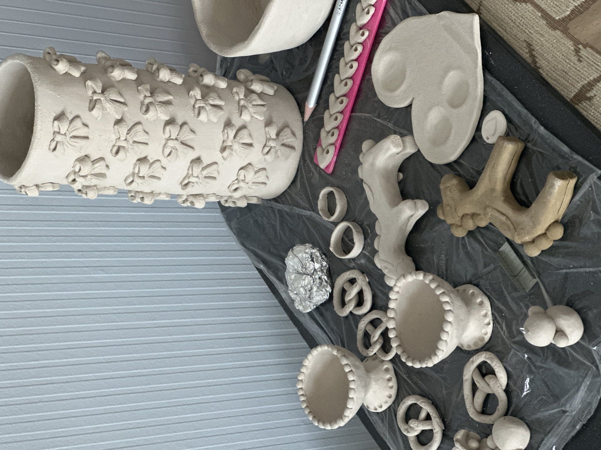 The image shows a collection of unfired clay objects arranged on a dark surface, likely a table covered with plastic sheeting. In the background, a light blue, vertically ribbed wall is visible.
The primary subject is a tall, cylindrical vase adorned with numerous small clay bows attached in a patterned arrangement. To the right of the vase, a portion of a bowl is visible.
Scattered across the surface are various clay creations:
* Two small pedestal bowls, one with a beaded rim.
* Several clay pretzels.
* Two clay rings.
* A clay animal figure, possibly a horse.
* A two-legged clay animal or figure, possibly a stylized horse.
* A heart-shaped clay object with three indentations.
* A small, round clay object with a hole, possibly a charm.
* A small, spherical clay figure resembling a bear.
* A crumpled piece of aluminum foil.
Other items present include a pink clay sculpting tool with multiple indentations, a silver pencil, and a small metal stapler. The objects appear to be in various stages of completion, suggesting a crafting or sculpting session. The scene does not contain any identifiable landmarks or contextual clues specific to Nevinnomyssk, Russia.