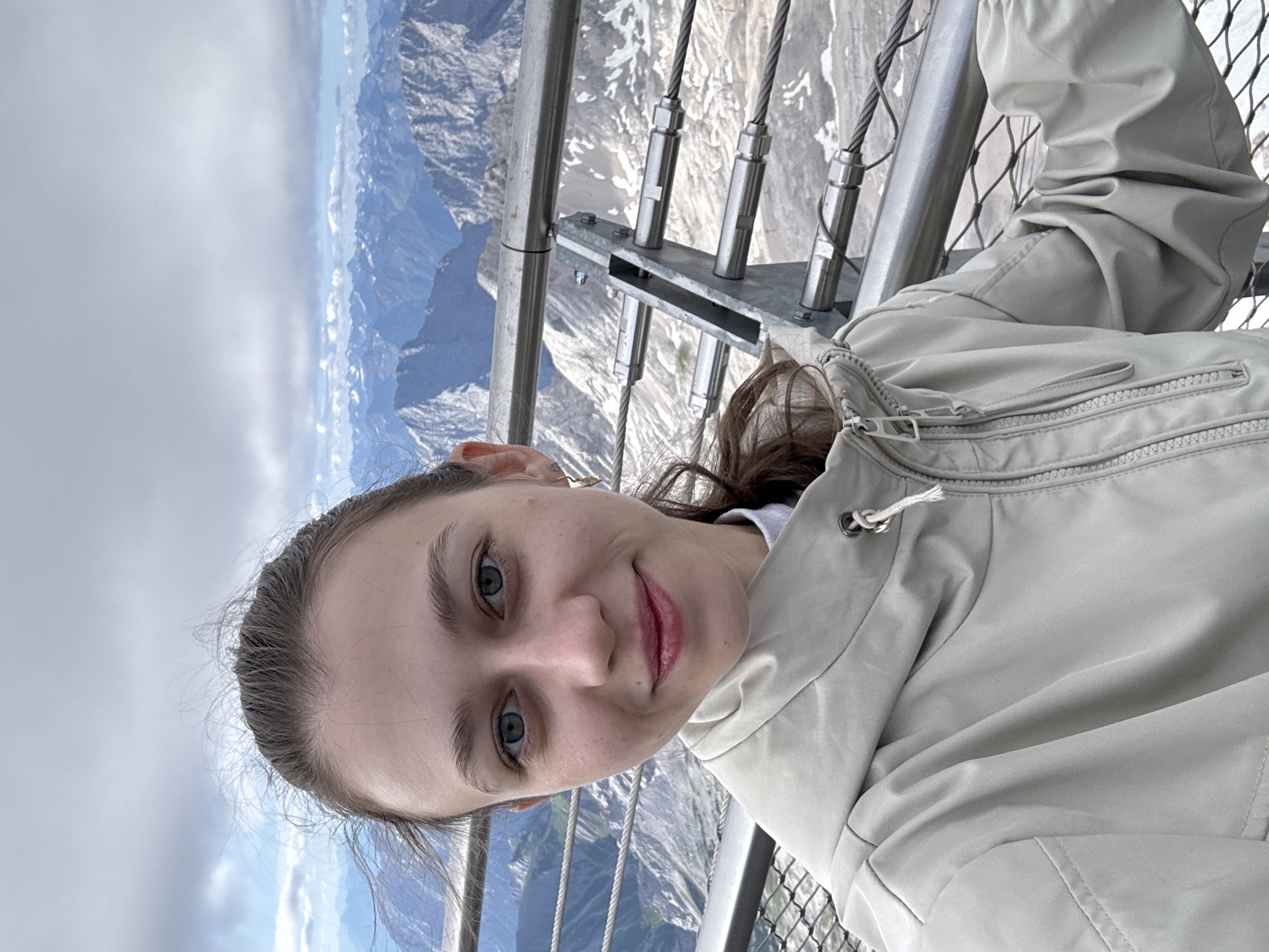 A young woman with brown hair pulled back and blue eyes smiles at the camera, taking a selfie. She is wearing a light-colored hooded jacket. Behind her, a metal railing and wire mesh fence are visible, with a vast mountain landscape stretching into the distance under a cloudy sky. The image appears to be taken from a high vantage point, possibly a viewing platform on a mountain. This scenery is characteristic of the Zugspitze, Germany's highest peak, located near the city of Garmisch-Partenkirchen.