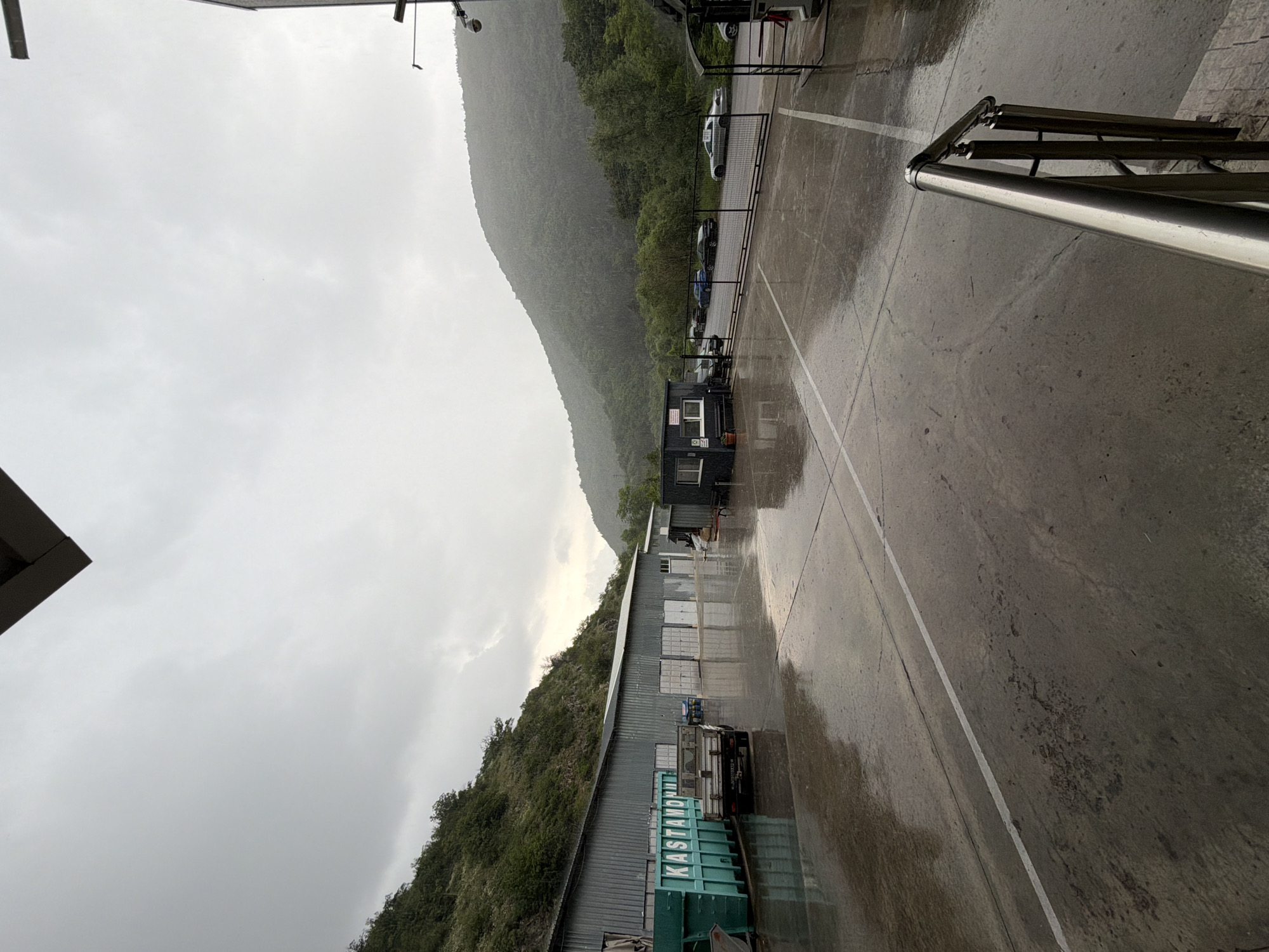 The image captures an outdoor scene dominated by natural landscape and industrial structures under an overcast sky. A large, steep, tree-covered mountain or hill occupies the left side of the frame, its verdant slopes receding into the distance. The sky above is entirely grey with thick clouds, indicating an overcast day.
In the foreground and middle ground, a wide paved area, possibly a road or parking lot, appears wet and reflective, suggesting recent or ongoing rain. White painted lines are visible on the asphalt. To the left of this paved area, a long, low-rise building with a corrugated metal roof and walls stretches across the scene, appearing to be an industrial facility or warehouse. Several rectangular openings or doors are visible along its side. A truck with a large, turquoise-colored container, bearing the white text "KASTAMONU", is parked next to this building.
Further down the paved area, past the main building, a smaller, dark-colored structure, possibly a guard booth or office, is situated. Several cars are parked beyond a metal fence that runs alongside the paved area, with more forested hills visible in the background. On the right side of the image, a portion of a shiny metal railing is visible in the foreground, suggesting the viewpoint is from an elevated platform or entrance.
No human activity is explicitly visible, rendering the scene quiet and static. The grey, cloudy sky and wet surfaces create a somewhat muted atmosphere. Based on the provided context, the location is Sliven, Bulgaria.
