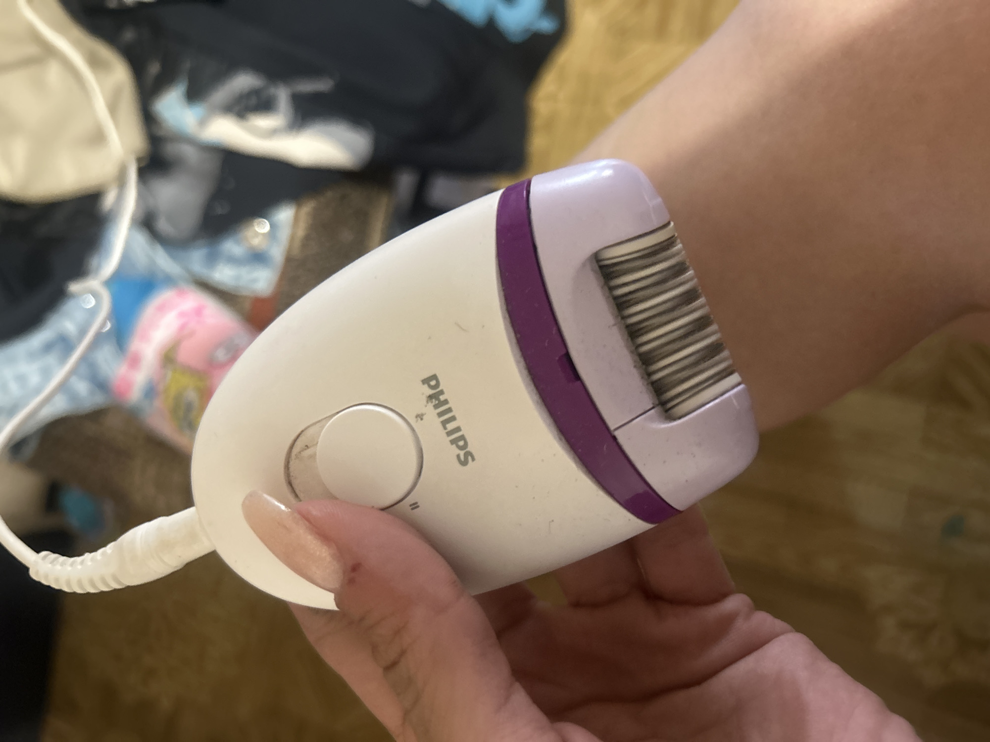 The image shows a close-up of a Philips epilator being held by a person's hand. The epilator is white with a purple trim and has a head with multiple metal tweezers. The person's thumb is resting on a circular button on the epilator. The background is blurred, but appears to be an indoor setting with some fabric items. There is no indication of the city of Perushtitsa, Bulgaria, or any specific urban environment.