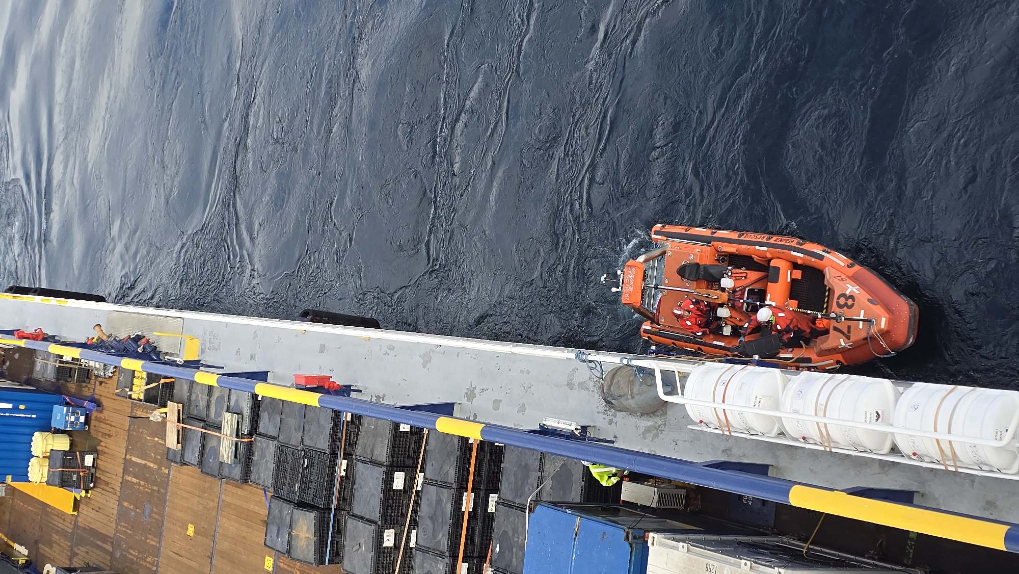 The image shows an orange inflatable rescue boat, labeled "ESKAY RESCUE," alongside a vessel. The boat is occupied by individuals in red and white protective gear. The vessel features a gray deck area with blue and yellow railings, various storage containers, and equipment. The surrounding environment is a body of water, with visible wave patterns. The location is Unknown, Unknown.