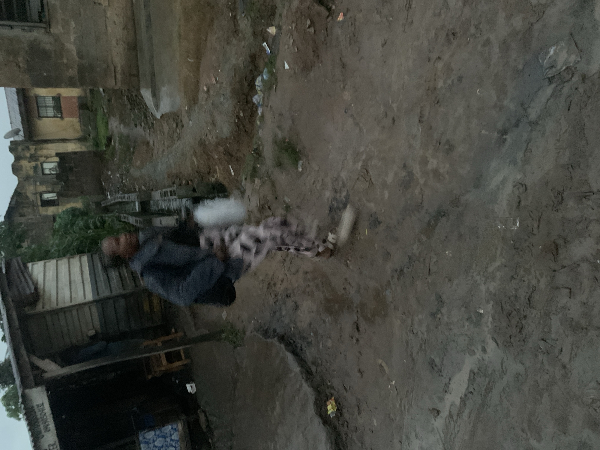 The image depicts a rural street scene in Shagamu, Nigeria, during what appears to be twilight or overcast conditions. The foreground is dominated by a muddy, uneven dirt road, showing signs of recent rain with puddles and damp patches. Debris, including plastic bags and wrappers, is scattered along the roadside.
In the mid-ground, a person is captured mid-stride, wearing a dark jacket and patterned trousers. They are carrying a white plastic bag. To the left, a makeshift stall or shop is visible, constructed with corrugated iron sheeting and wooden planks. A chair and a blue and white patterned fabric are seen inside the stall.
The background reveals a cluster of buildings, some appearing weathered and in disrepair, with exposed brickwork and decaying plaster. One building has a satellite dish mounted on its roof. Vegetation is sparse, with some greenery visible between the structures. The overall atmosphere suggests a modest, possibly impoverished, urban or peri-urban environment.