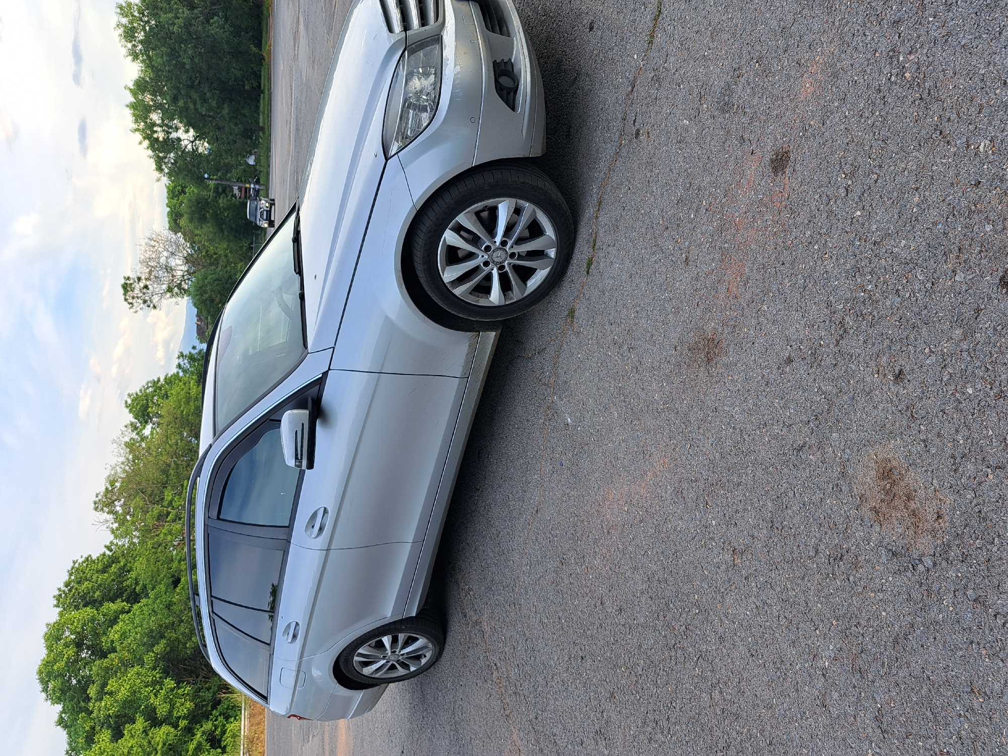 A silver Mercedes-Benz C-Class Estate (S204) is parked on an asphalt surface. The car is facing the right side of the frame, with its front end slightly angled towards the camera. The vehicle has tinted windows and silver alloy wheels. In the background, there are trees and a cloudy sky. To the right, a dark SUV is visible in the distance. The location is identified as the parking lot of a roadside restaurant in Lokorsko, Bulgaria. The scene appears to be an outdoor setting during daylight.