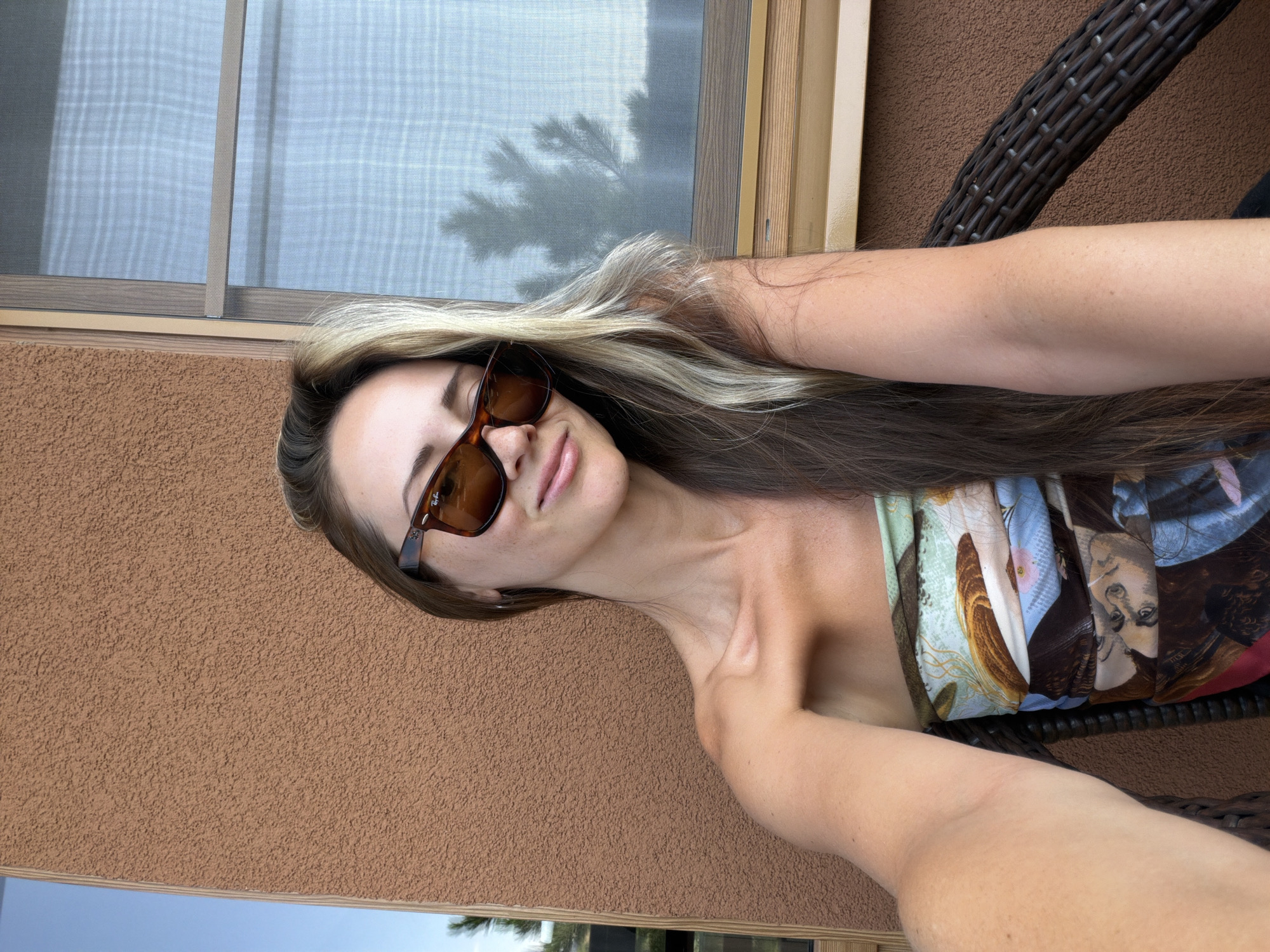 In the city of Kamyshin, Russia, a young woman with long, blonde-streaked brown hair, wearing sunglasses and a patterned strapless top, is captured in a selfie. She is seated in a dark wicker chair, outdoors, against a textured brown wall. To her right, a window with a screen is visible, showing the blurry silhouette of a tree. The lighting suggests it is daytime.