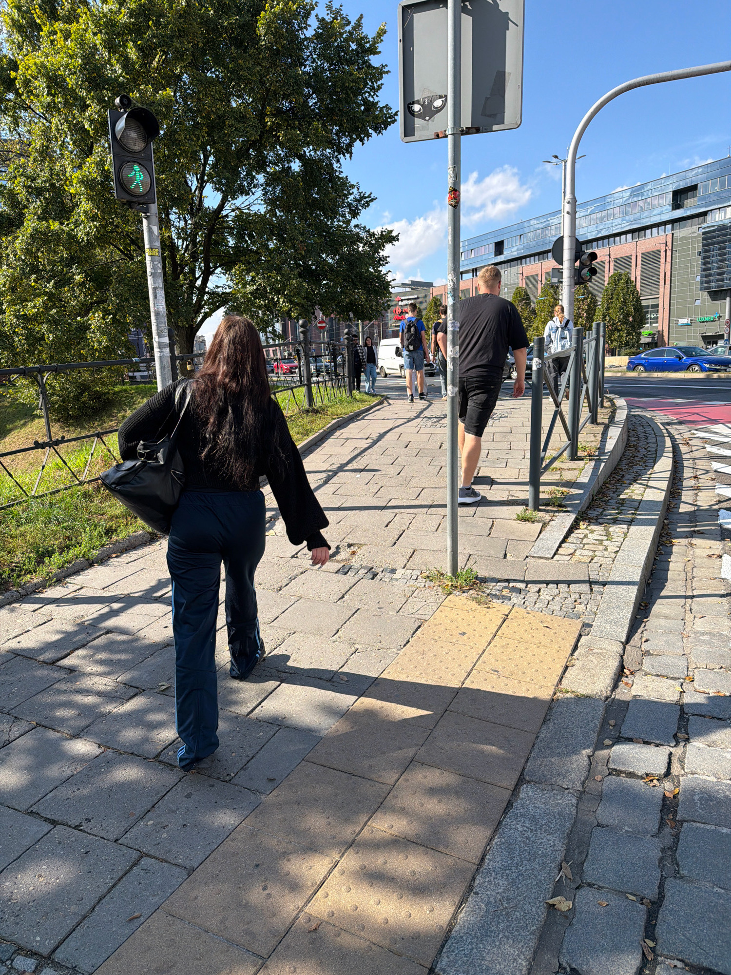 The image captures a sunny day on a city street in Wrocław, Poland, likely near a pedestrian crossing. In the foreground, a woman with long dark hair is walking away from the camera, wearing a black knitted sweater and dark blue track pants with light blue stripes. She is carrying a large black shoulder bag. The sidewalk is paved with rectangular concrete tiles and bordered by cobblestones. To her left, a metal fence separates the sidewalk from a grassy area. A traffic light with a green pedestrian signal is visible on a pole. In the background, several people are walking, and a blue car is driving on the road. Buildings with modern architecture line the opposite side of the street. The scene suggests a normal day in an urban environment.