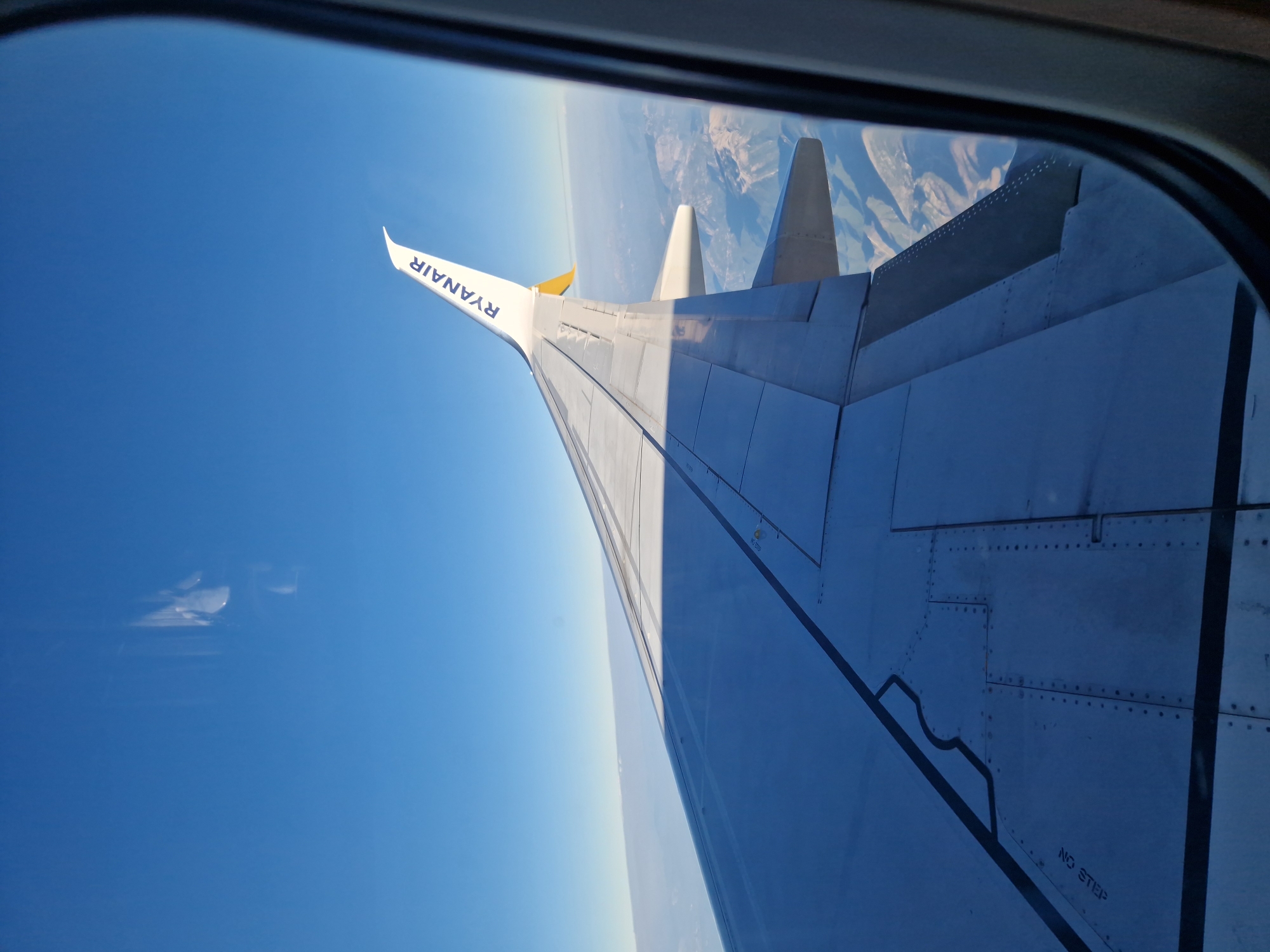 An aerial view from an airplane window shows a portion of a Ryanair airplane wing extending into the frame, with the "RYANAIR" logo visible on the wingtip. The wing is predominantly white with black accents and rivets. Below the wing, a vast landscape of mountains and valleys is visible, partially obscured by a layer of clouds. The sky is a clear, bright blue. The perspective suggests the airplane is in flight, likely over a mountainous region. No specific location in the city of Pandalejmon, Albania, can be identified from this image.