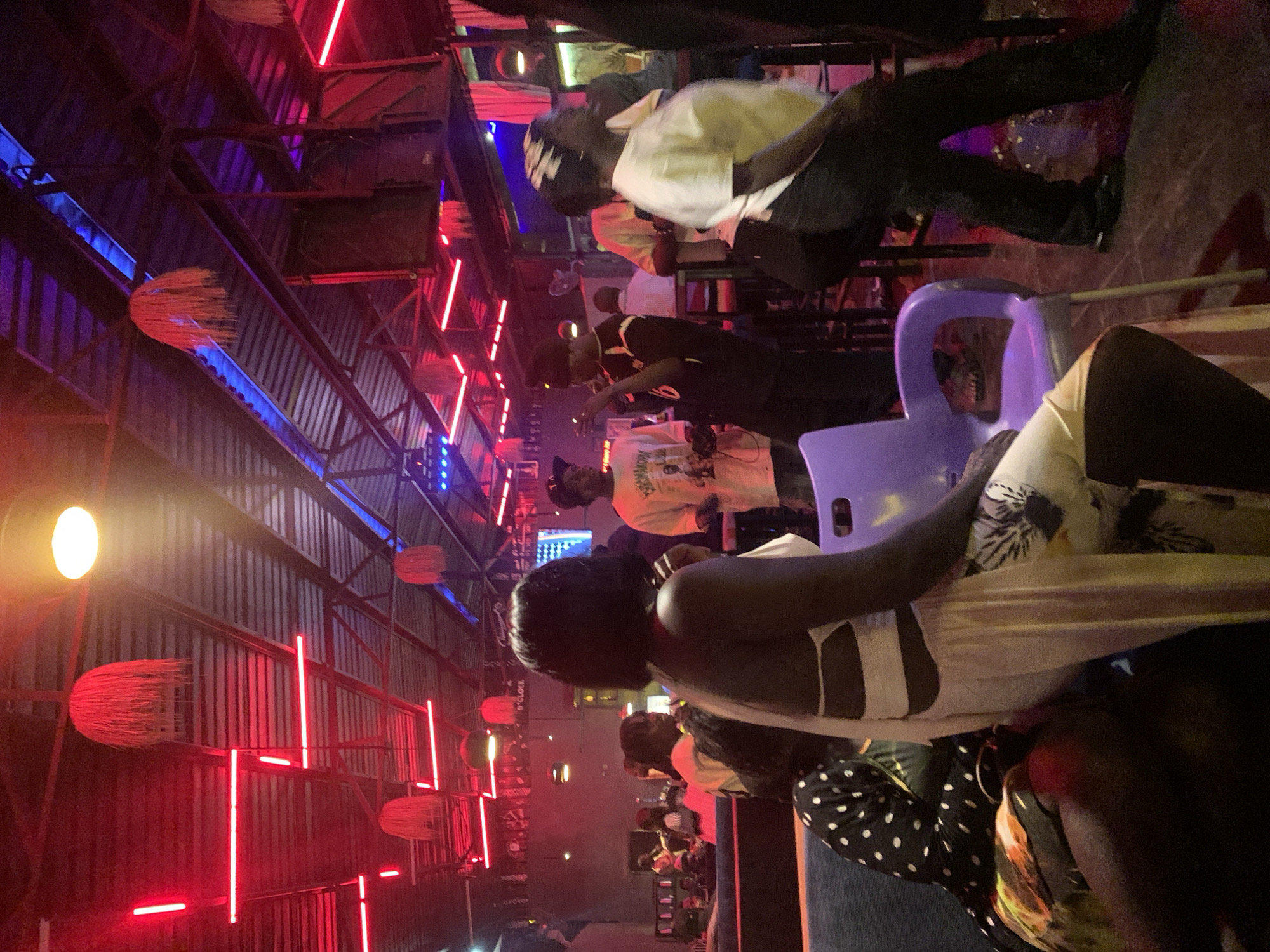 The image depicts an indoor scene at a venue on Rukuba Road, Nigeria, characterized by a vibrant, low-light atmosphere illuminated by neon red and blue lighting. The ceiling is a corrugated metal structure with exposed beams, from which several woven, spherical light fixtures hang, resembling rustic chandeliers. Neon red LED strips are installed along the ceiling and walls, casting a dominant glow. In the foreground, a person is seated on a purple plastic chair, facing away from the camera and towards the right side of the frame. They are wearing a light-colored, possibly off-white, flowing garment with cutouts at the waist and patterned fabric on the legs. To the left of this seated person, another individual is partially visible, wearing a black and white polka dot top and a floral patterned skirt.
In the mid-ground, several other individuals are present, appearing to be patrons or staff. One person in a white t-shirt with graphics stands near a bar or counter area. Another person in a black t-shirt with white lettering is talking or gesturing. The background reveals more people, seating arrangements, and possibly a screen or stage area, suggesting a social gathering, entertainment venue, or restaurant. The floor is dark, with some reflections from the lighting. The overall impression is one of a lively and modern social establishment.