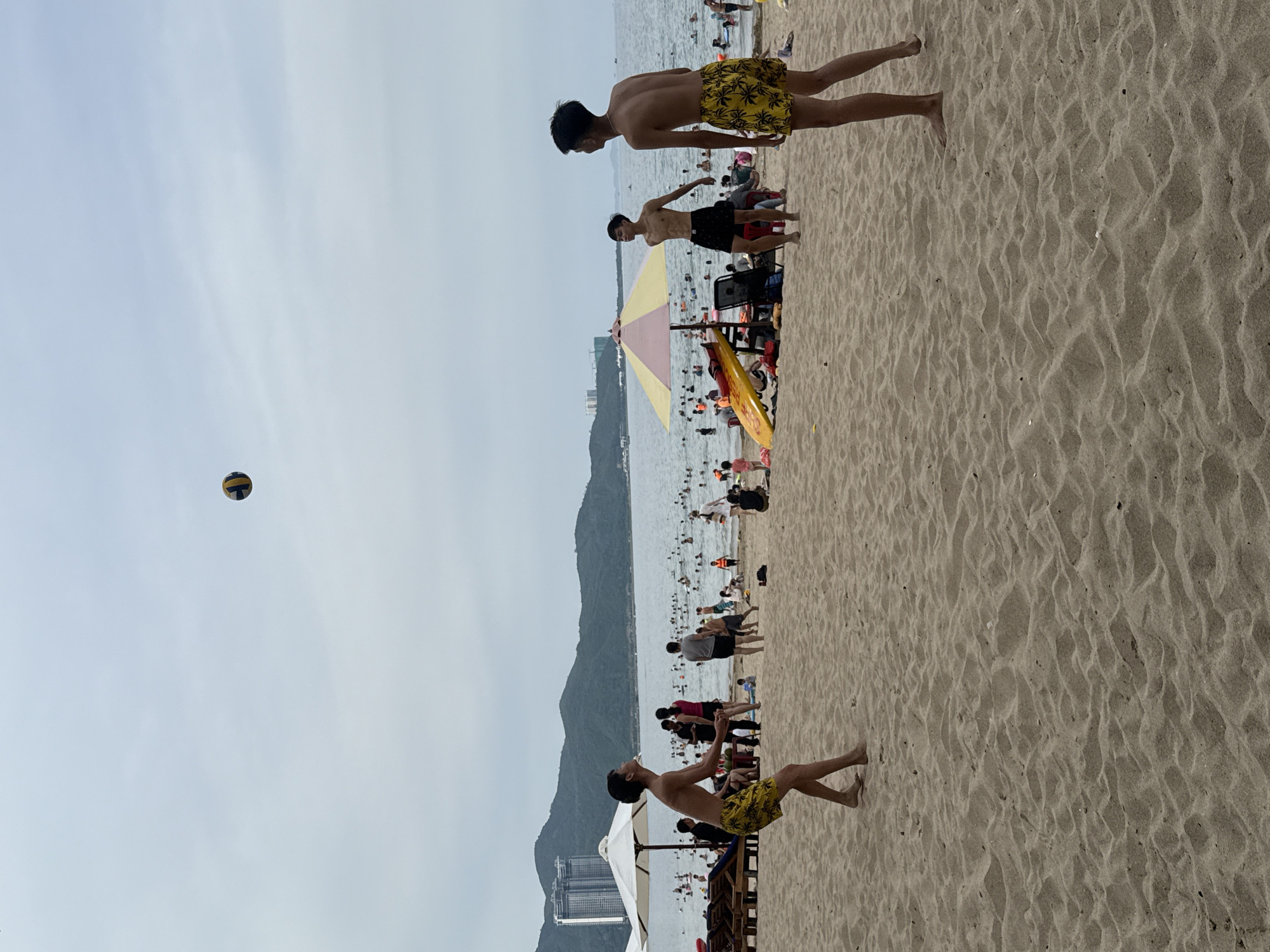 An overcast day at a crowded beach in Nha Trang, Vietnam, showcases several individuals engaged in a game of beach volleyball. In the foreground, two young men stand on the sand, one mid-serve with a volleyball in the air above him, and the other watching the play. In the mid-ground, another young man in black shorts stands near a yellow and pink umbrella, observing the game. Numerous beachgoers populate the water and sand, with lounge chairs and umbrellas set up on the beach. In the distance, a mountainous landscape forms the backdrop to the bustling scene, with a few buildings visible on the slopes.